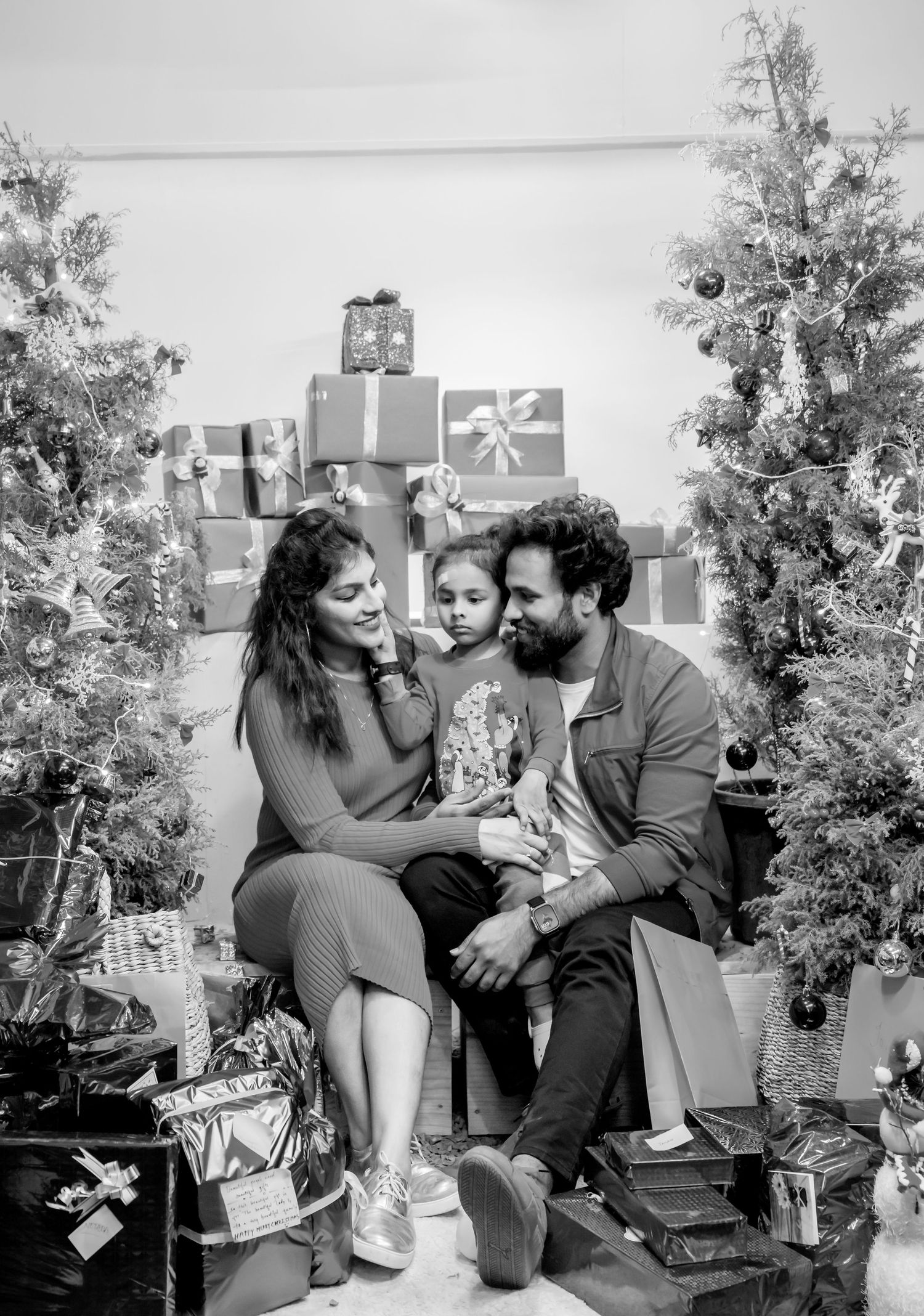 Black and white family Christmas photo with parents and child surrounded by decorated trees and wrapped gifts.