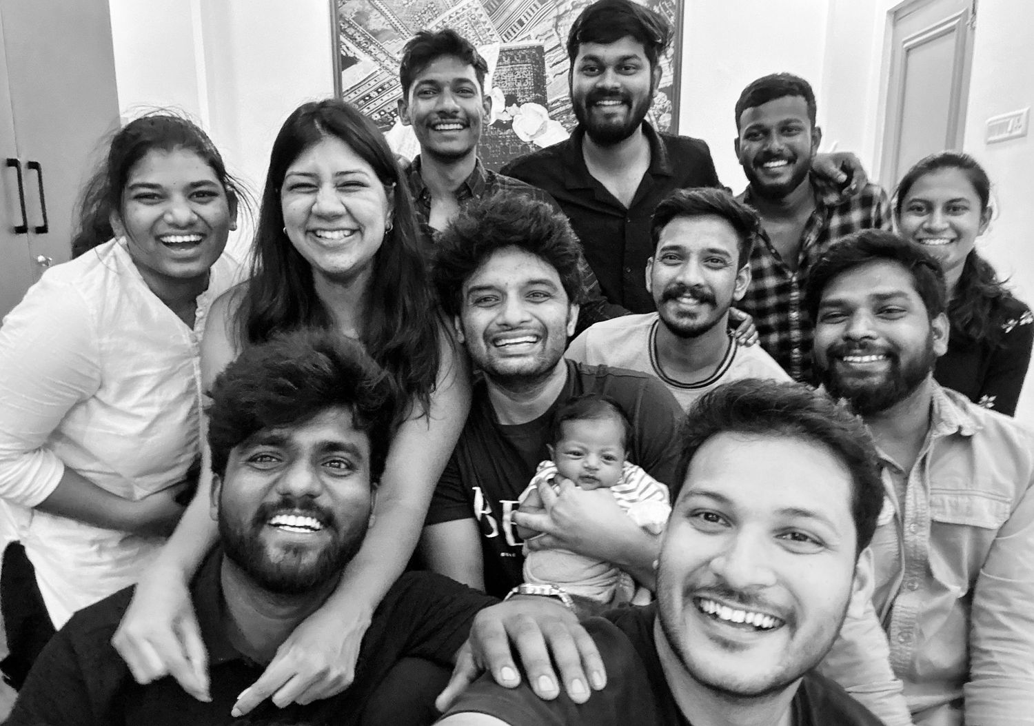 Black and white group photo of smiling friends posing together indoors, one person holding a baby.