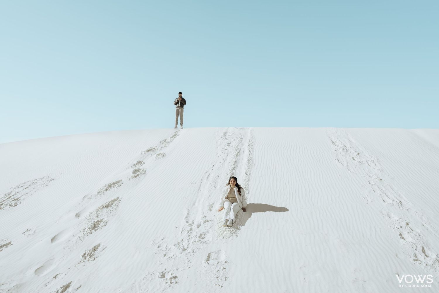 Couple on white sand dunes, woman in white dress walking down slope, man standing at top, clear blue sky.