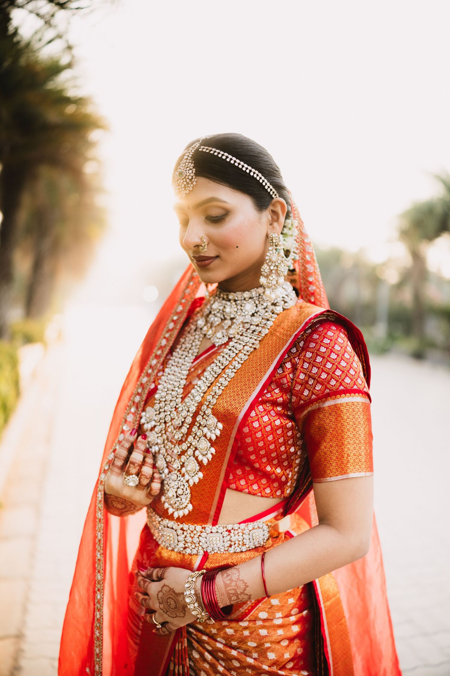 Indian bride in red and orange saree with layered pearl jewelry and maang tikka poses outdoors in golden light.