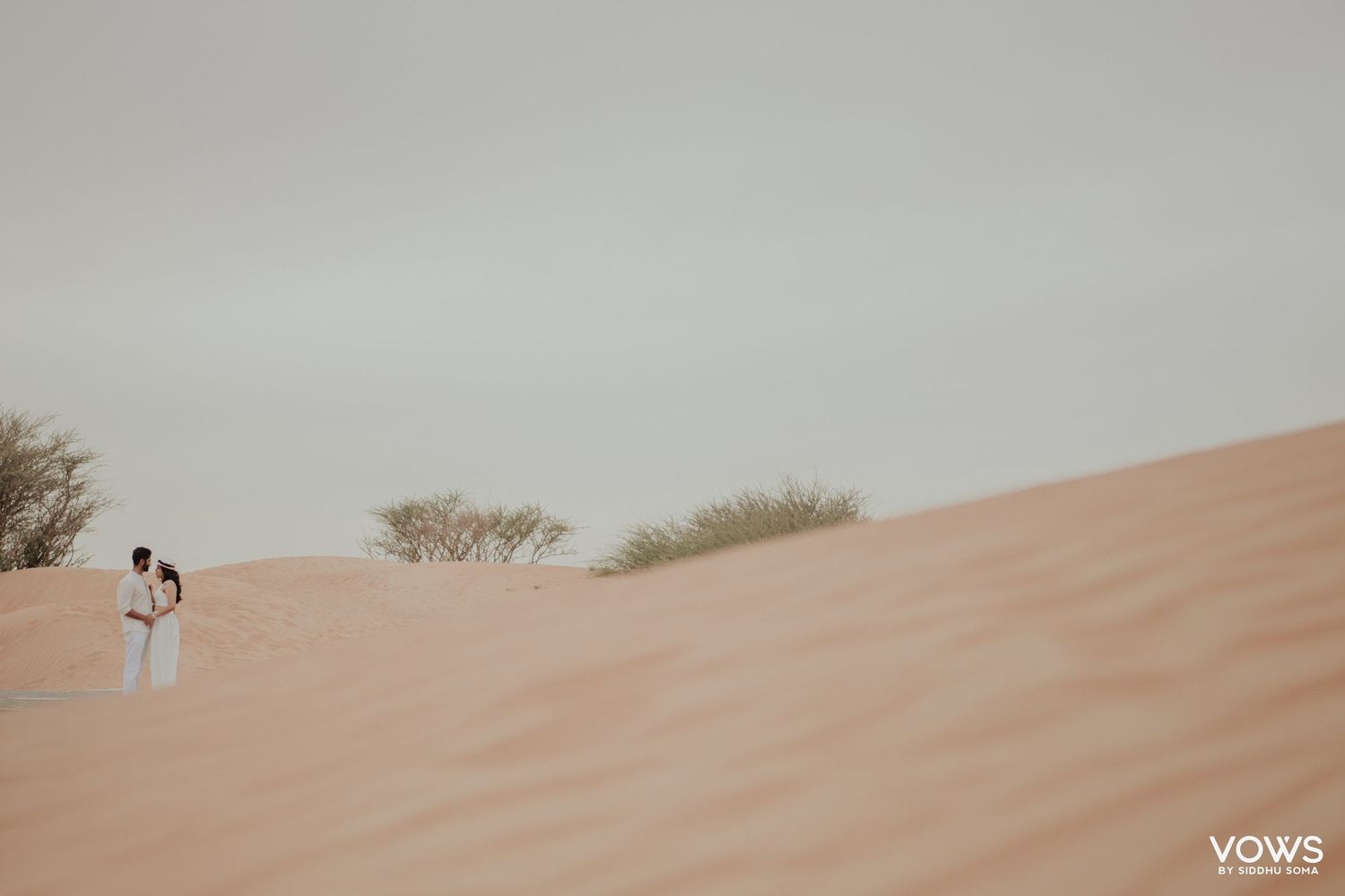 Couple in white embracing on a vast sandy desert dune with sparse green shrubs and a soft gray sky.