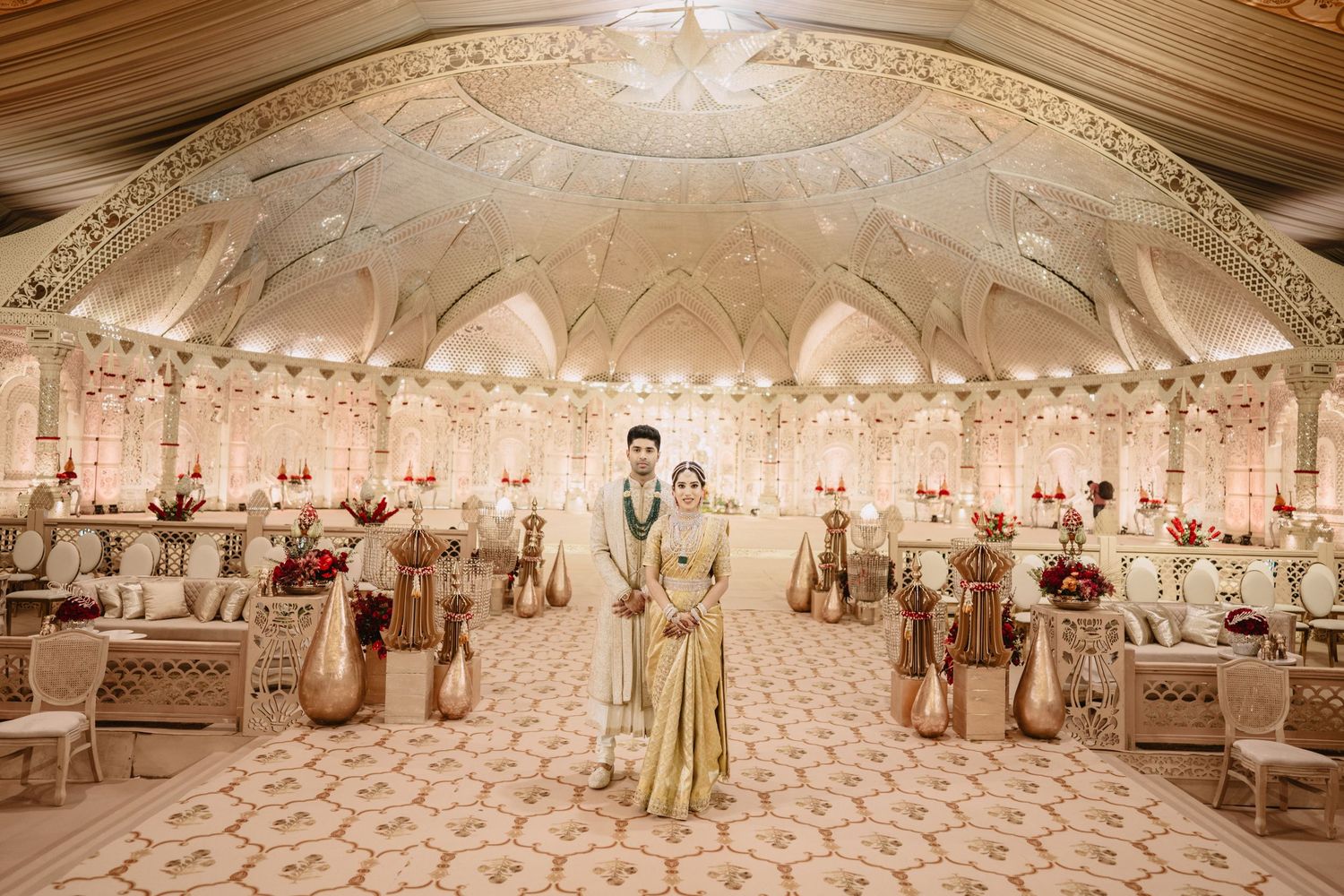 Bride and groom stand in ornate gold wedding reception hall with floral decor and elegant white arched ceiling.