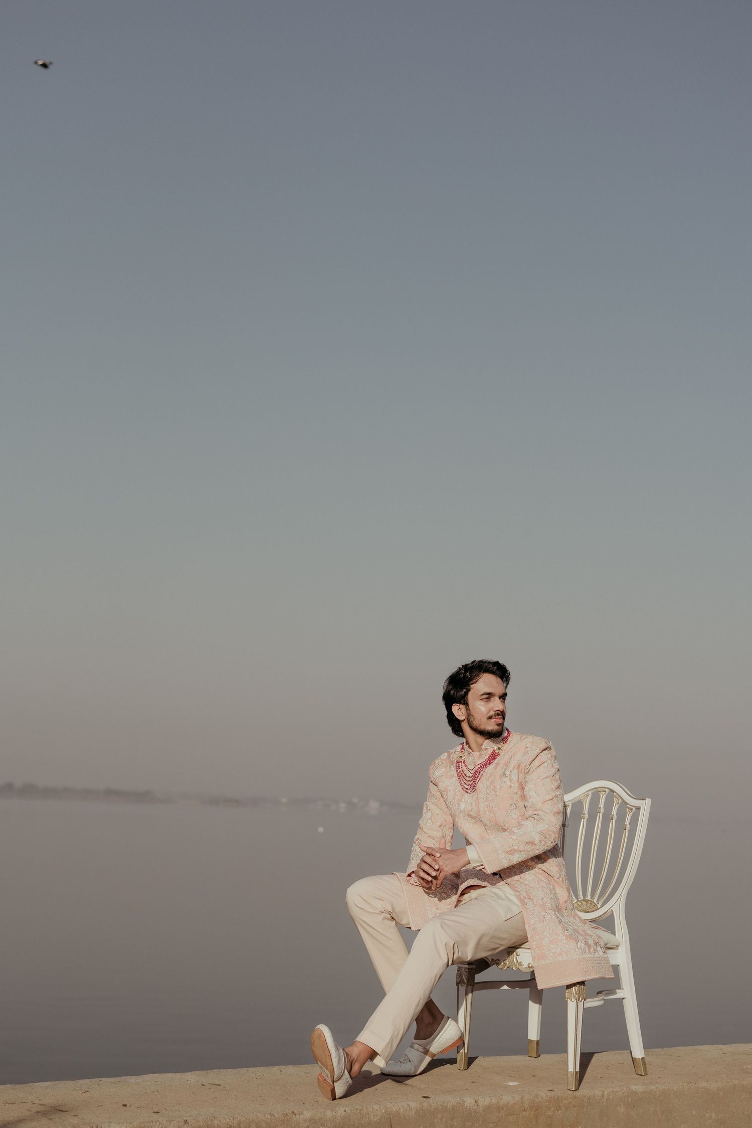 Groom in ivory sherwani sits on white chair by misty riverside in serene outdoor portrait.