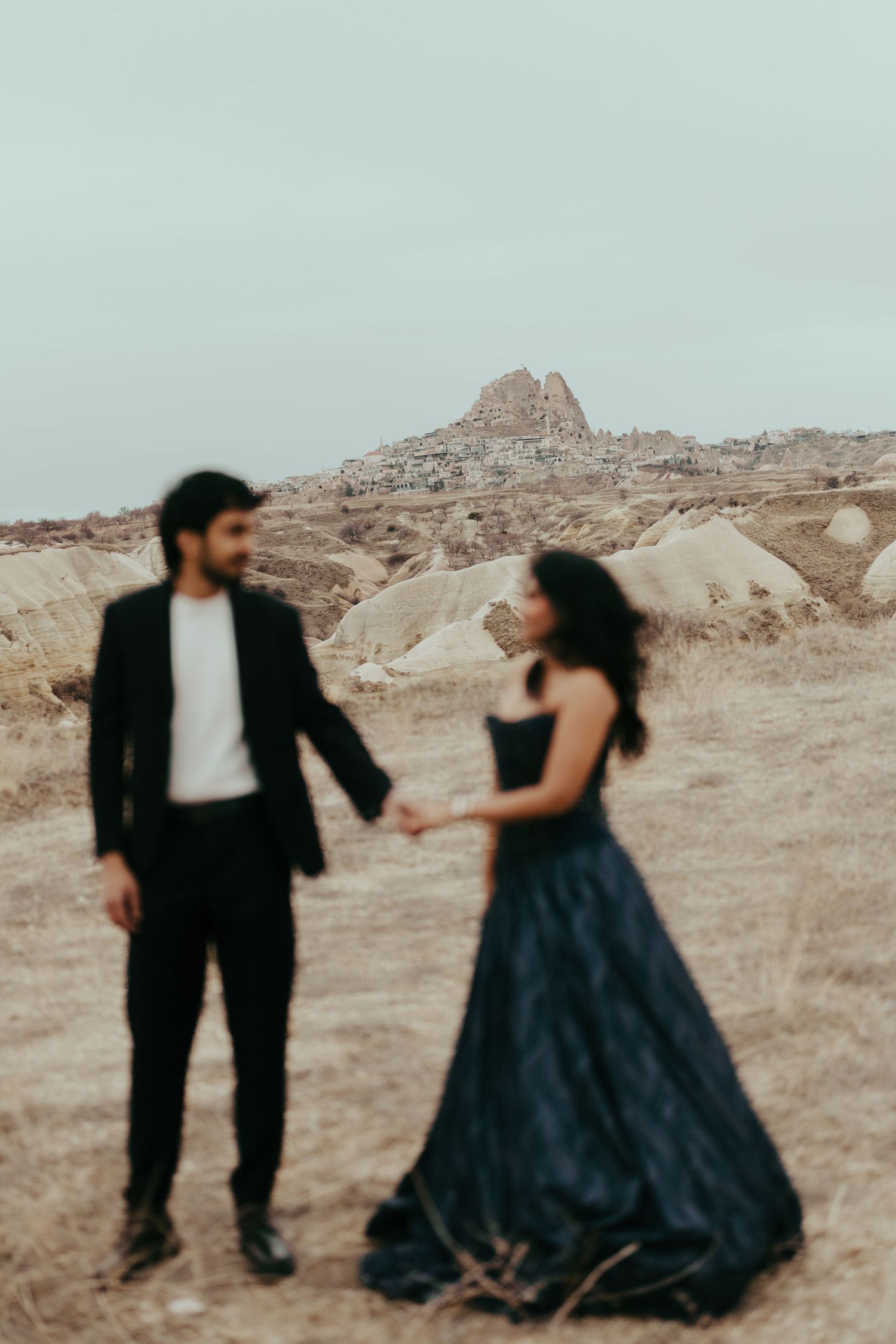 Couple holding hands in formal attire with dramatic Cappadocia rock formations in the background.