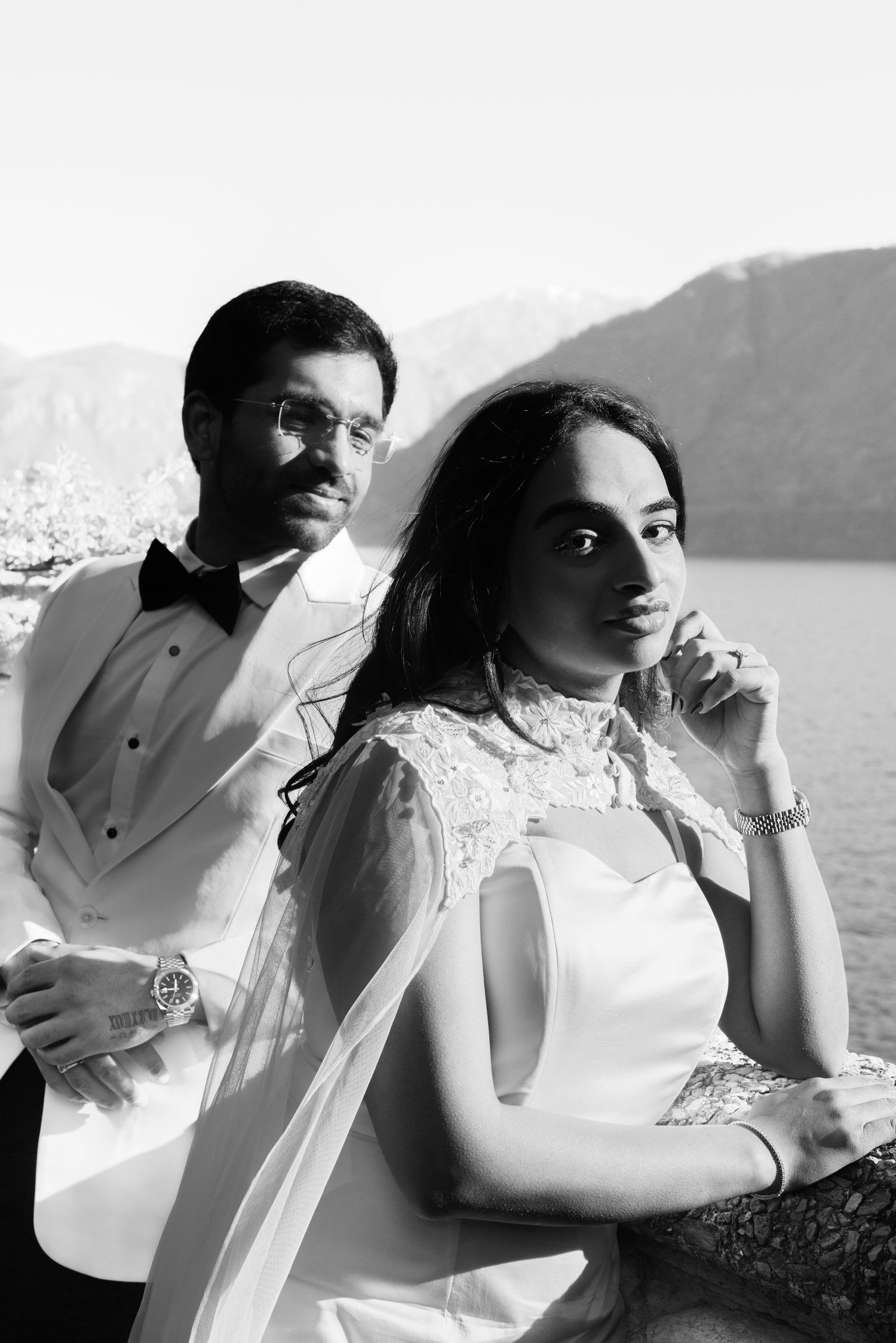 Elegant couple in white wedding attire pose by a mountain lake in a classic black and white portrait photo.