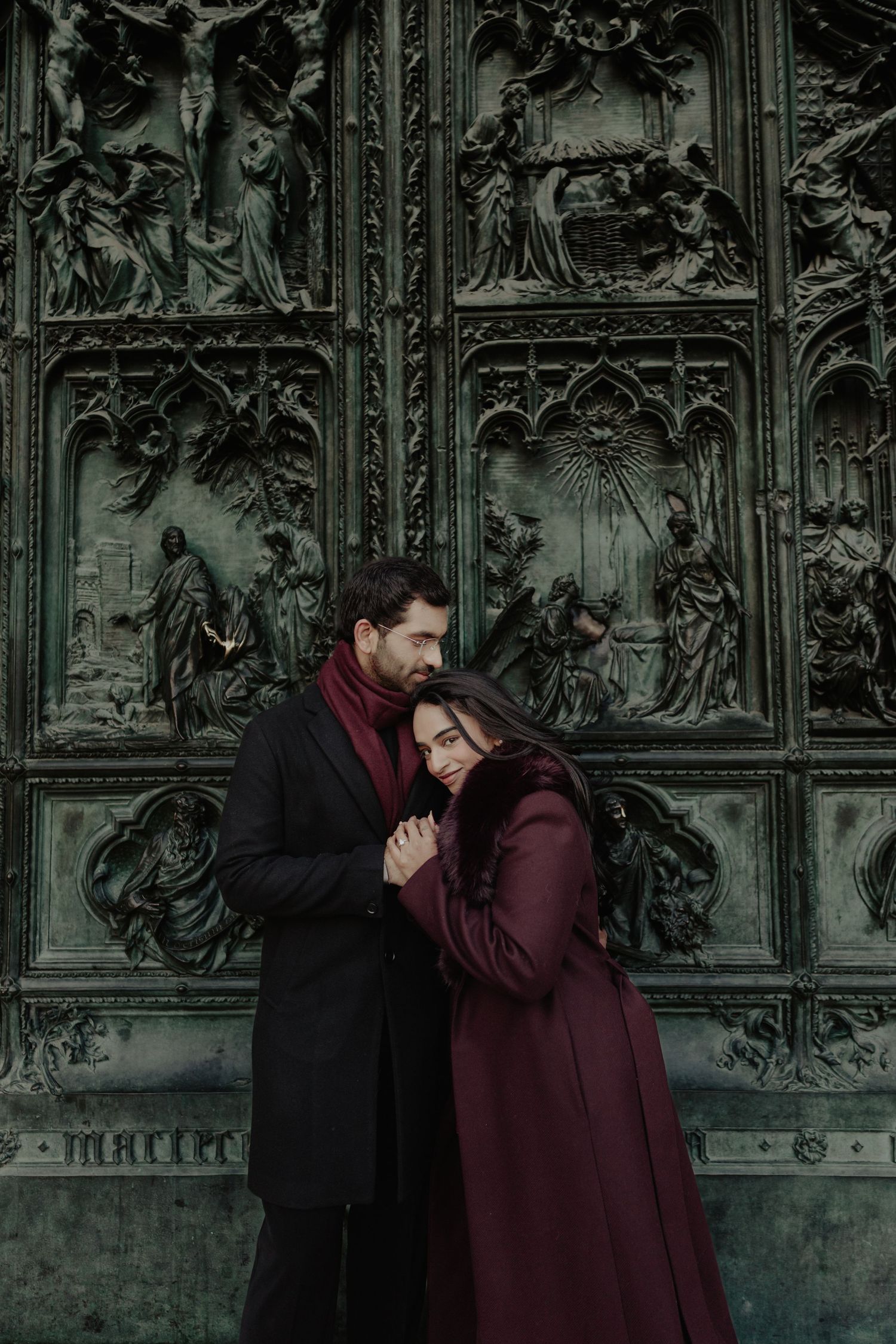 Romantic couple embracing in front of ornate bronze cathedral doors with religious relief sculptures.