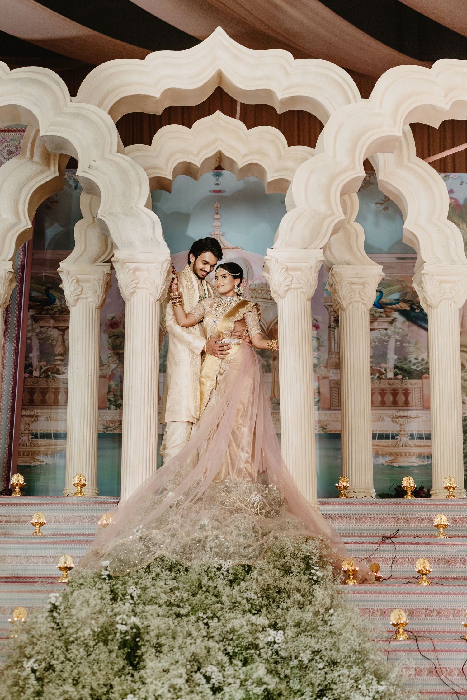 Bride and groom posing under ornate white Mughal arches surrounded by white floral arrangements.