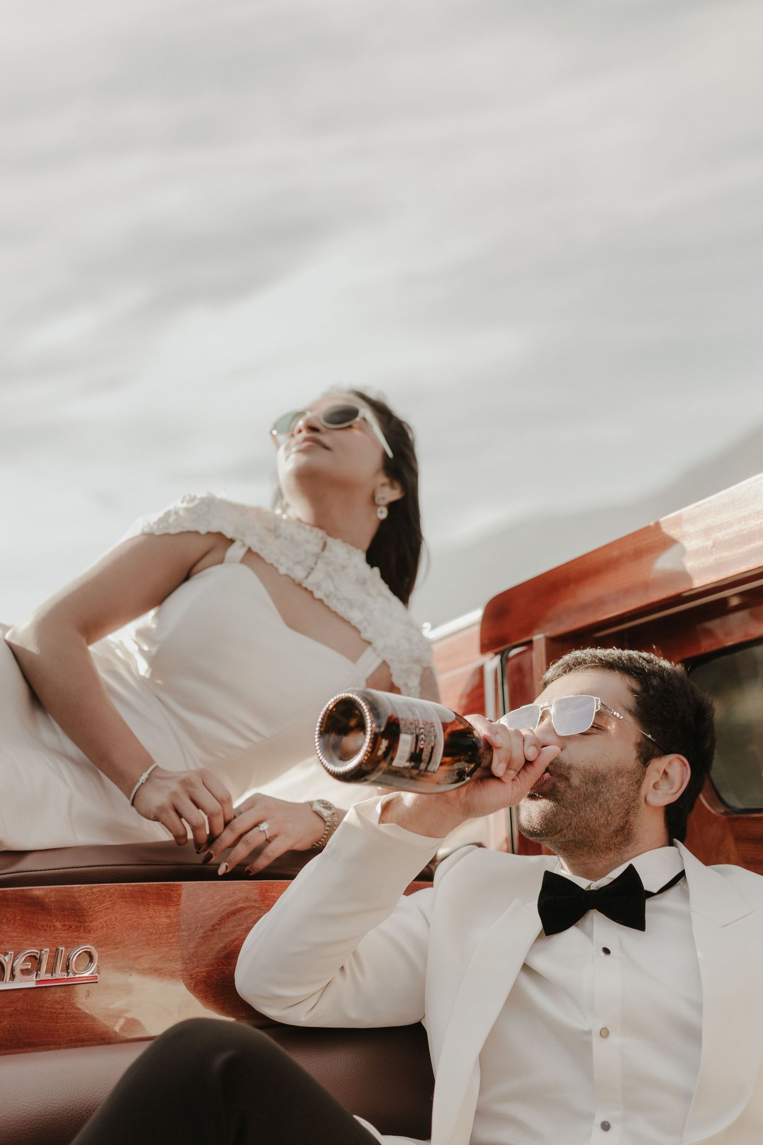 Stylish wedding couple on vintage boat, groom in tuxedo drinking champagne while bride poses in white gown.