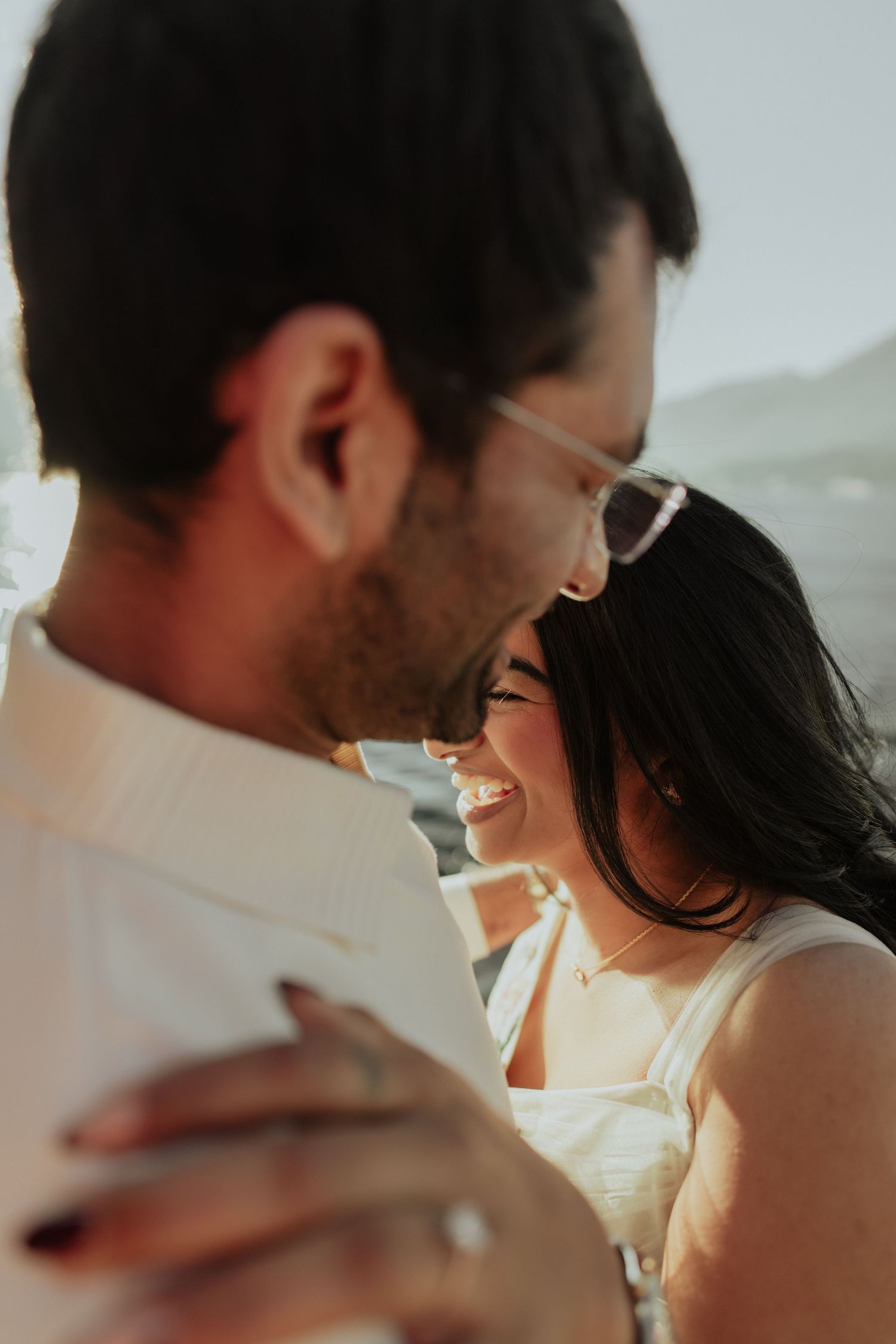 A smiling couple shares a romantic embrace outdoors, bathed in warm golden sunlight near a scenic waterfront.