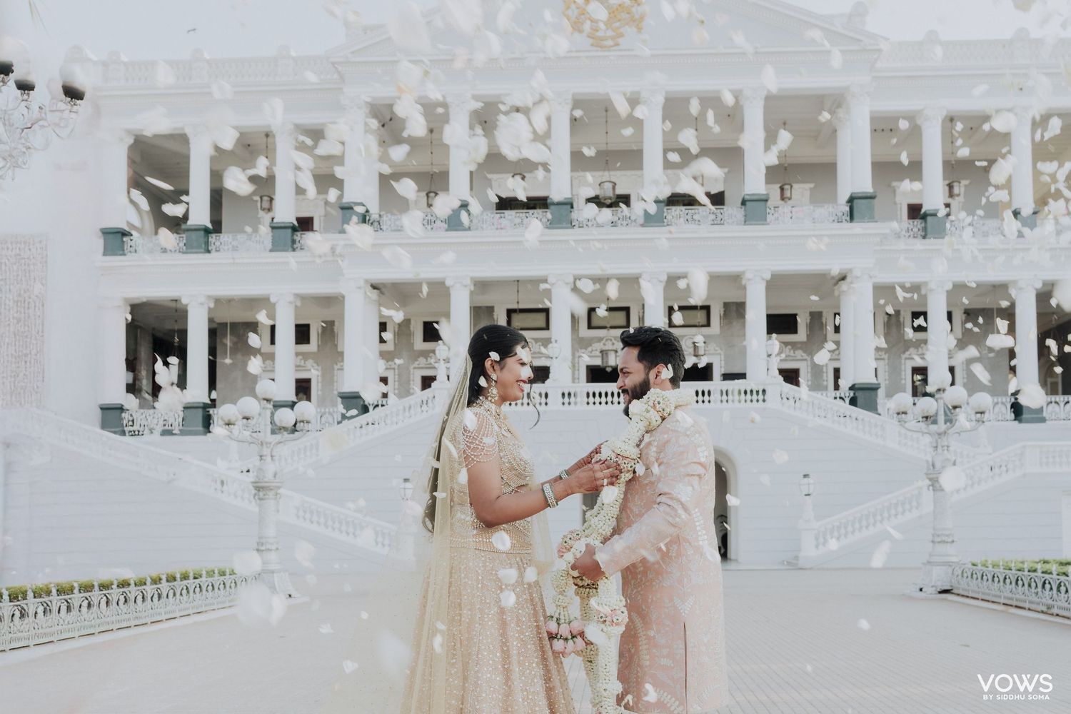 Bride and groom exchange vows at elegant white wedding venue with floral confetti falling around them.