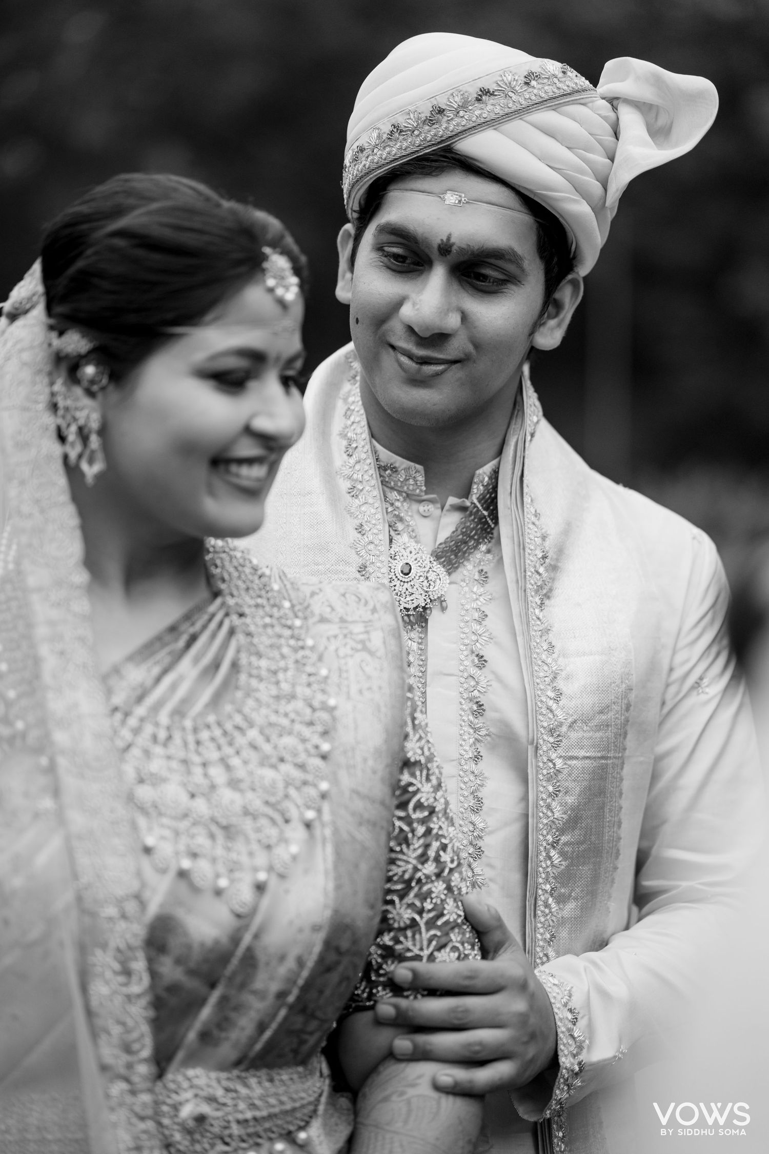 Black and white portrait of a smiling bride and groom in traditional Indian wedding attire, with ornate jewelry and a embroidered turban.