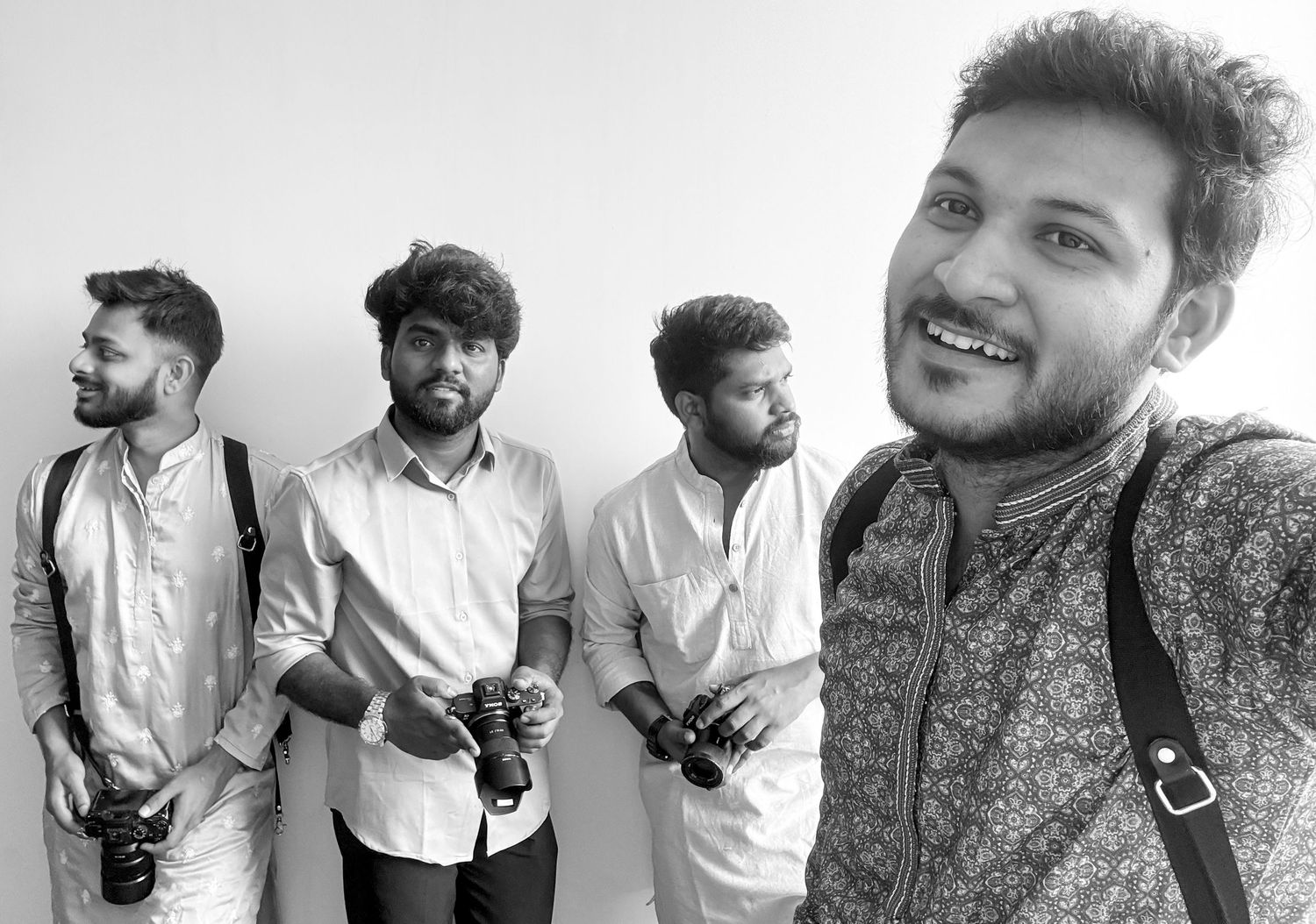 Black and white selfie of four young men with cameras, smiling outdoors in casual shirts.
