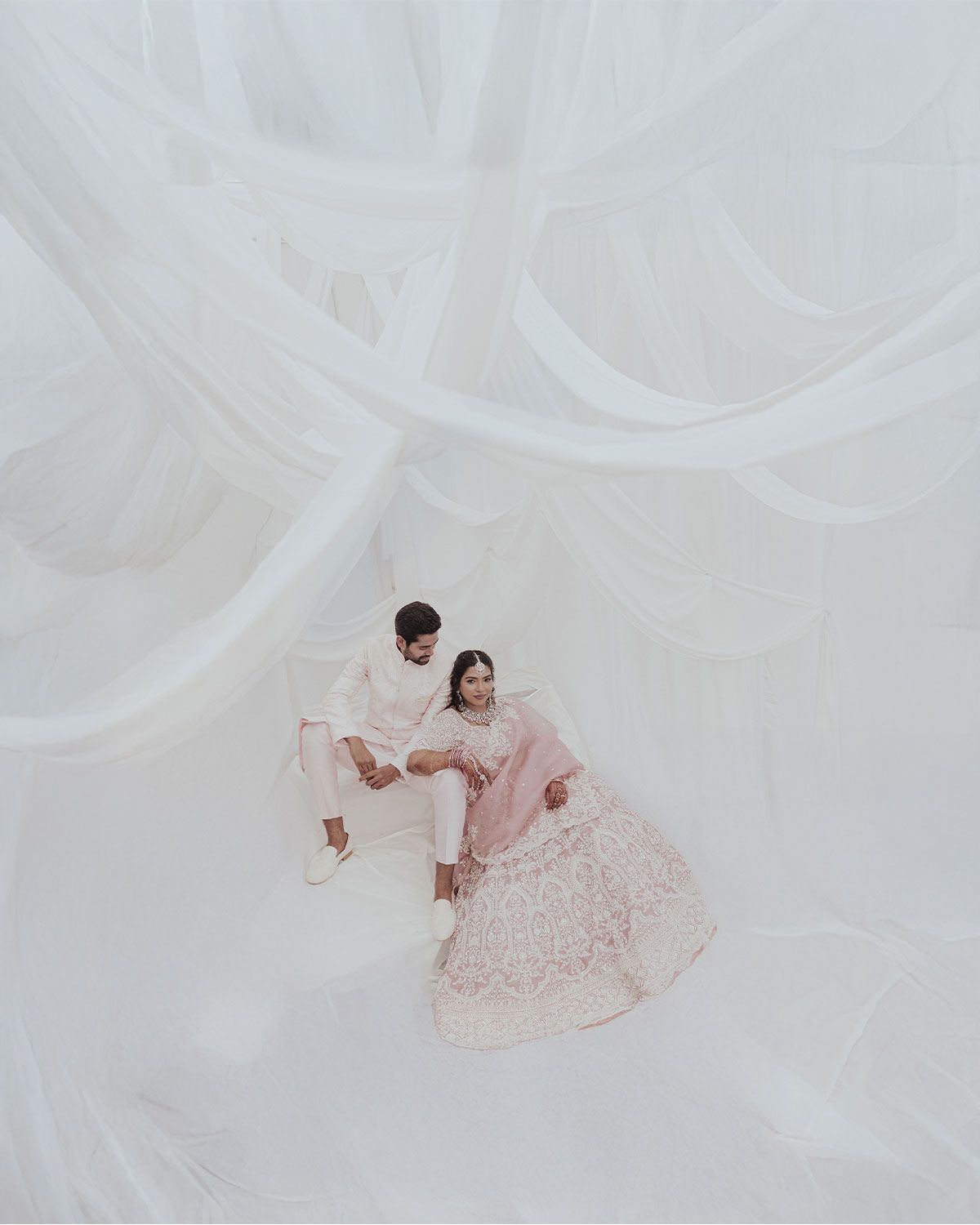 Couple in traditional South Asian wedding attire sitting together under flowing white drapes, bride in pink lehenga.