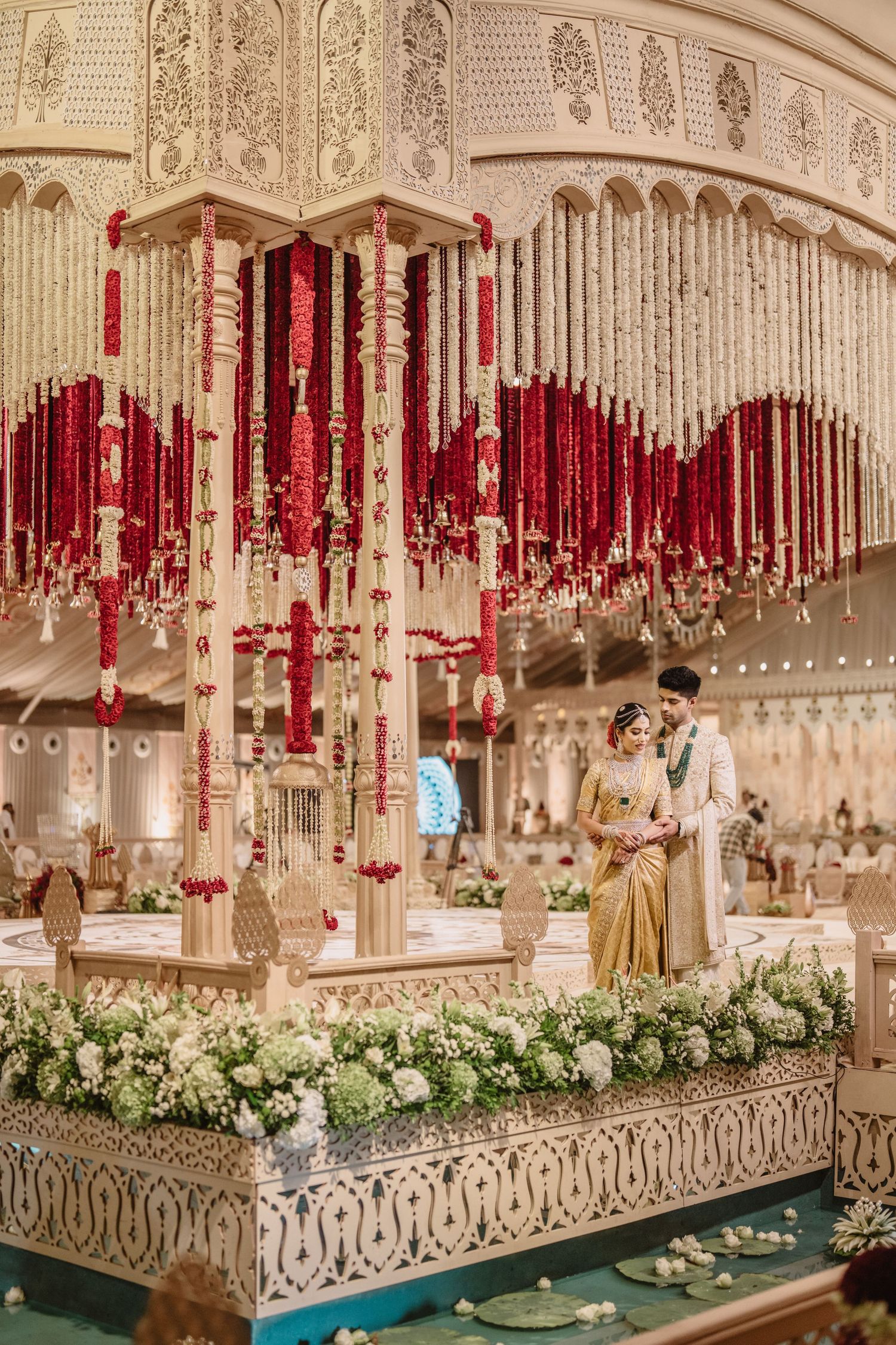 Indian wedding couple standing under an ornate mandap adorned with red and white floral garlands at a grand ceremony.