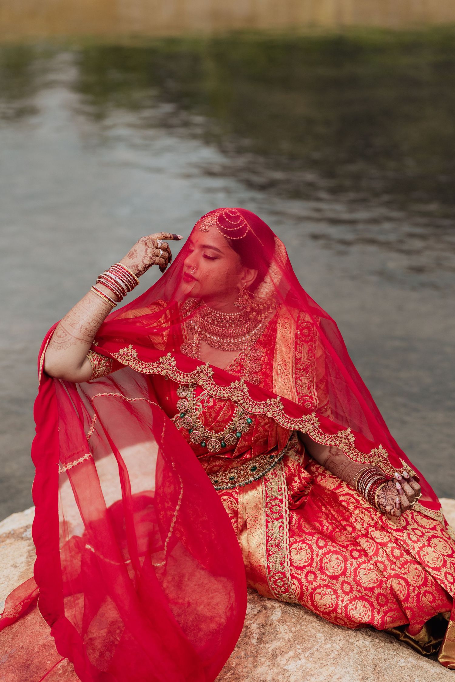 A bride in a red lehenga and dupatta sits gracefully by a lake, adorned with gold jewelry and mehndi.