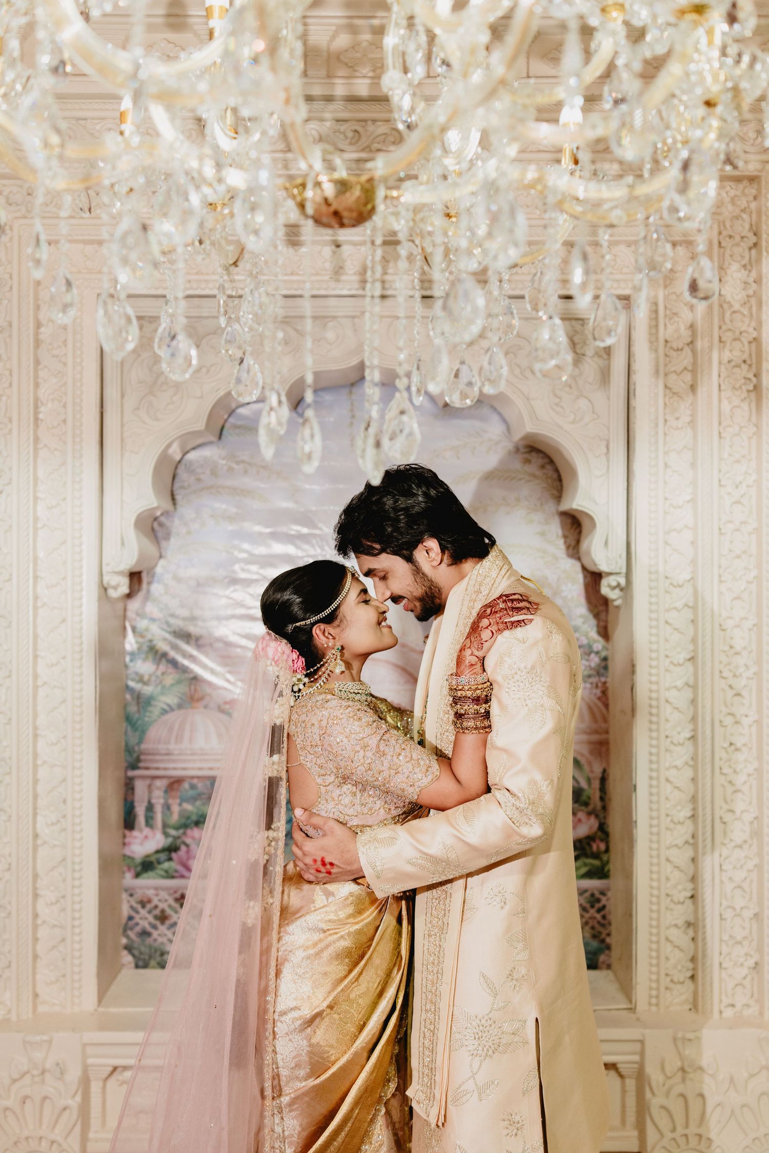 Indian bride and groom in gold wedding attire embracing under crystal chandelier in ornate white venue.