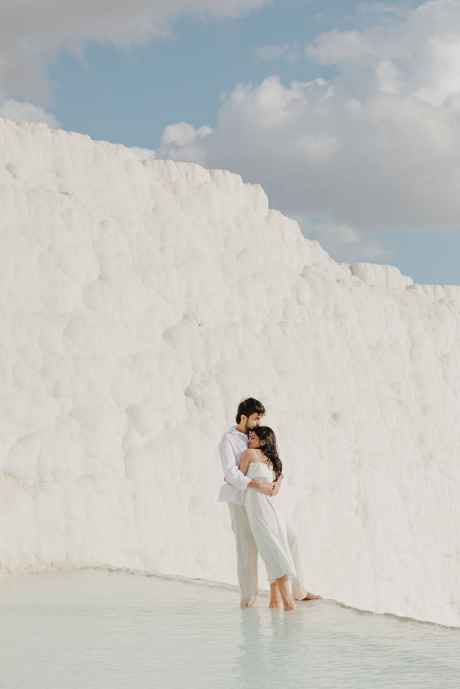A couple embraces barefoot in shallow water against the stunning white travertine terraces of Pamukkale, Turkey.