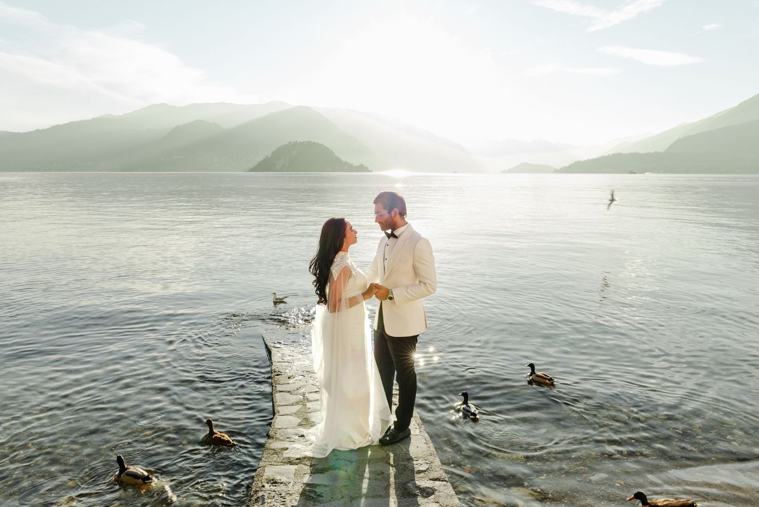 Bride and groom stand on a lakeside dock surrounded by ducks, with misty mountains and golden sunlight in the background.