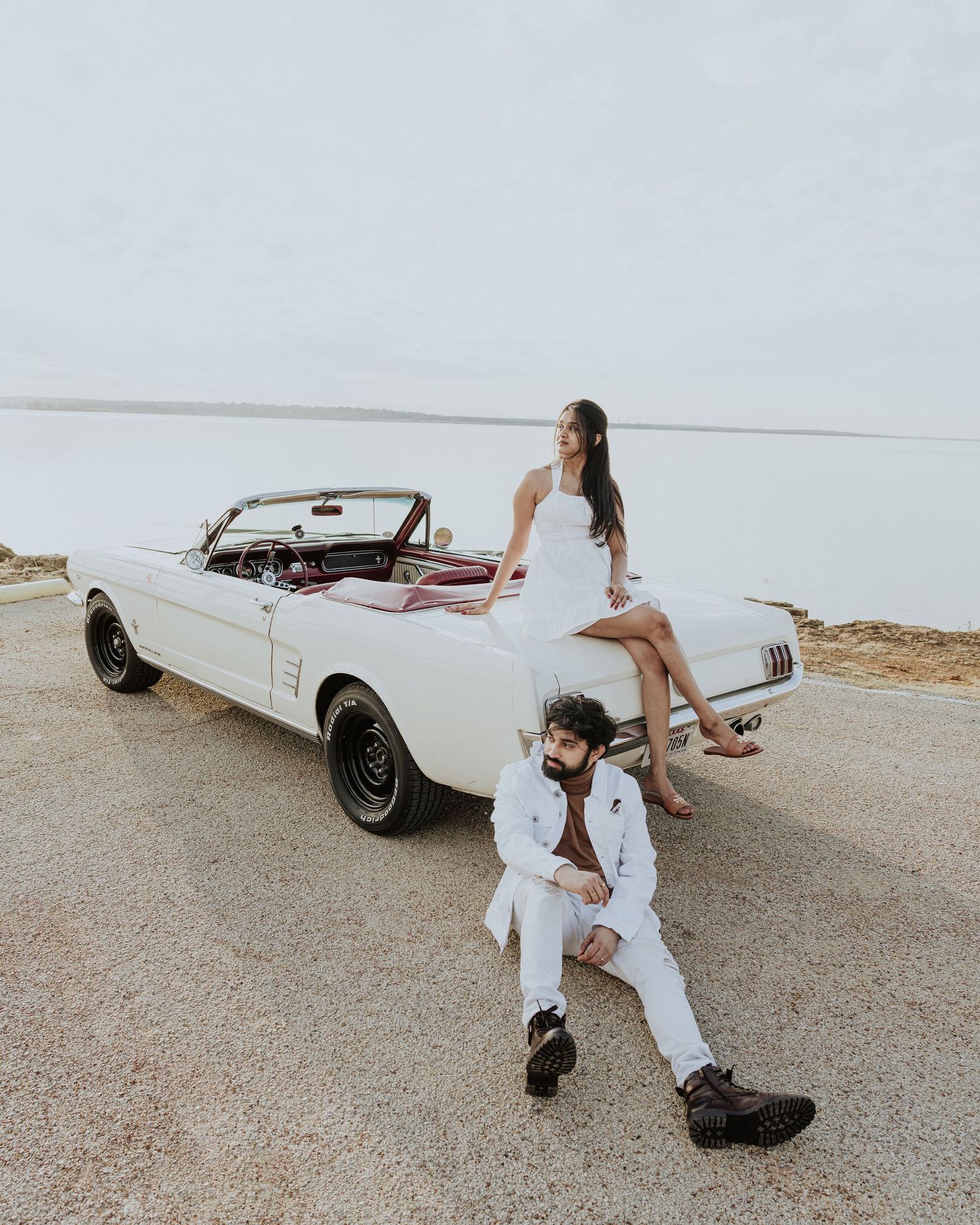 Bride sits atop a vintage white Ford Mustang convertible while groom leans against it on a scenic road.