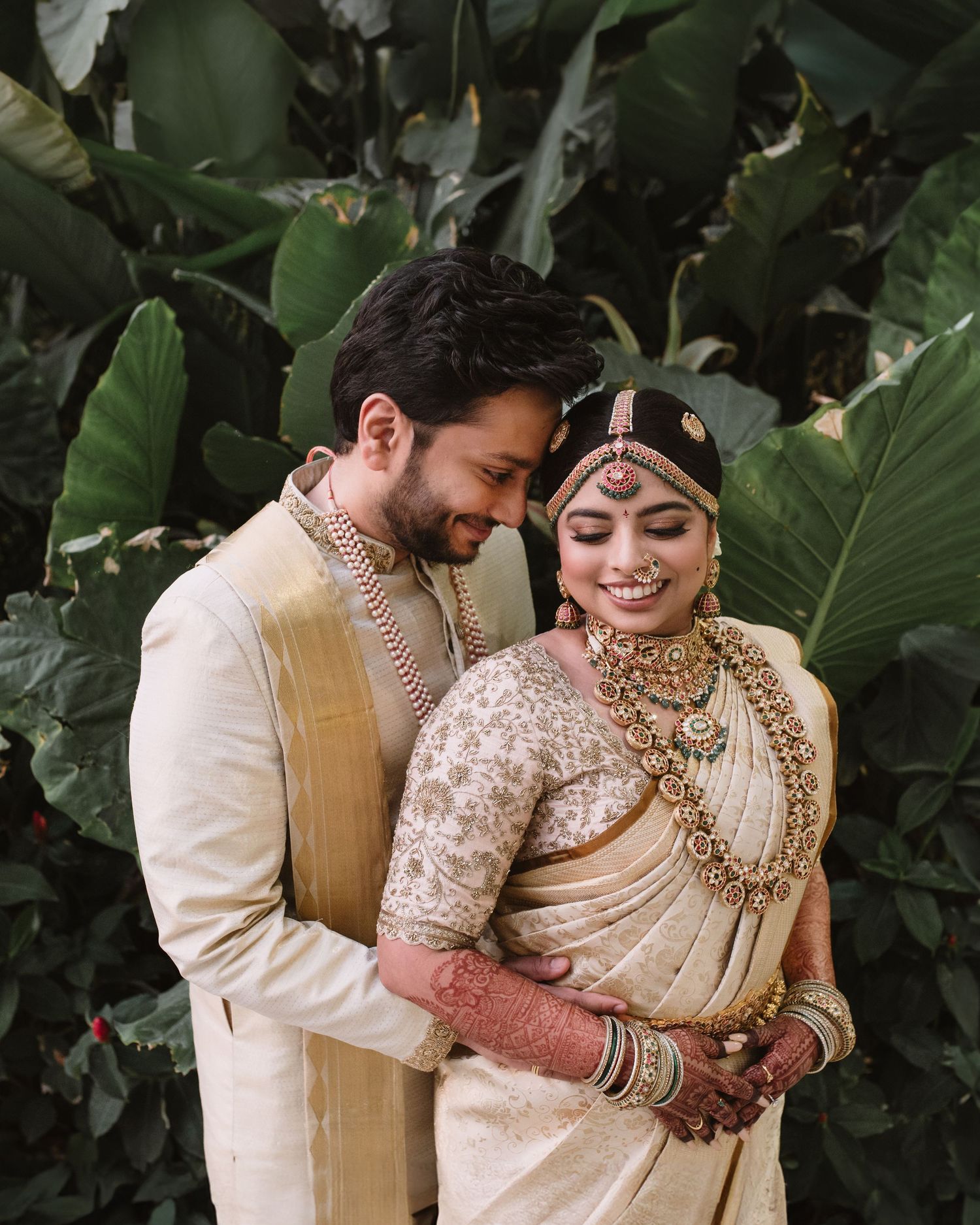 Indian bride and groom in traditional wedding attire, posing together against lush tropical green leaves backdrop.