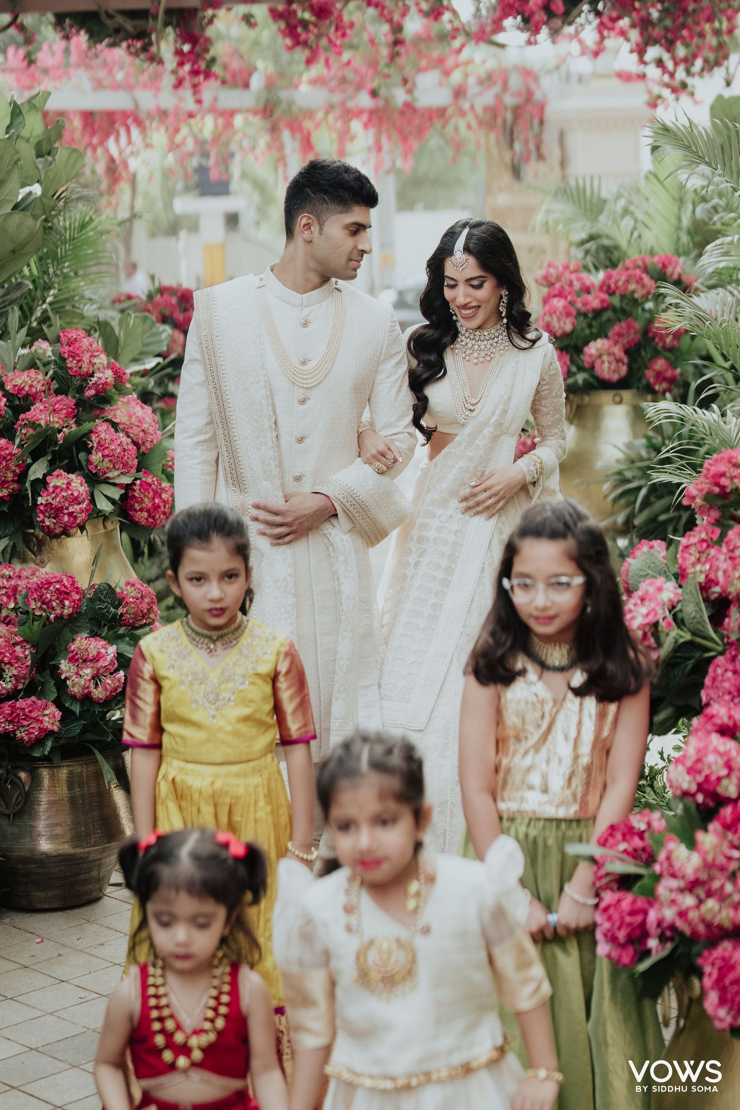 Elegant couple in white wedding attire surrounded by pink flowers and children in colorful traditional Indian outfits.