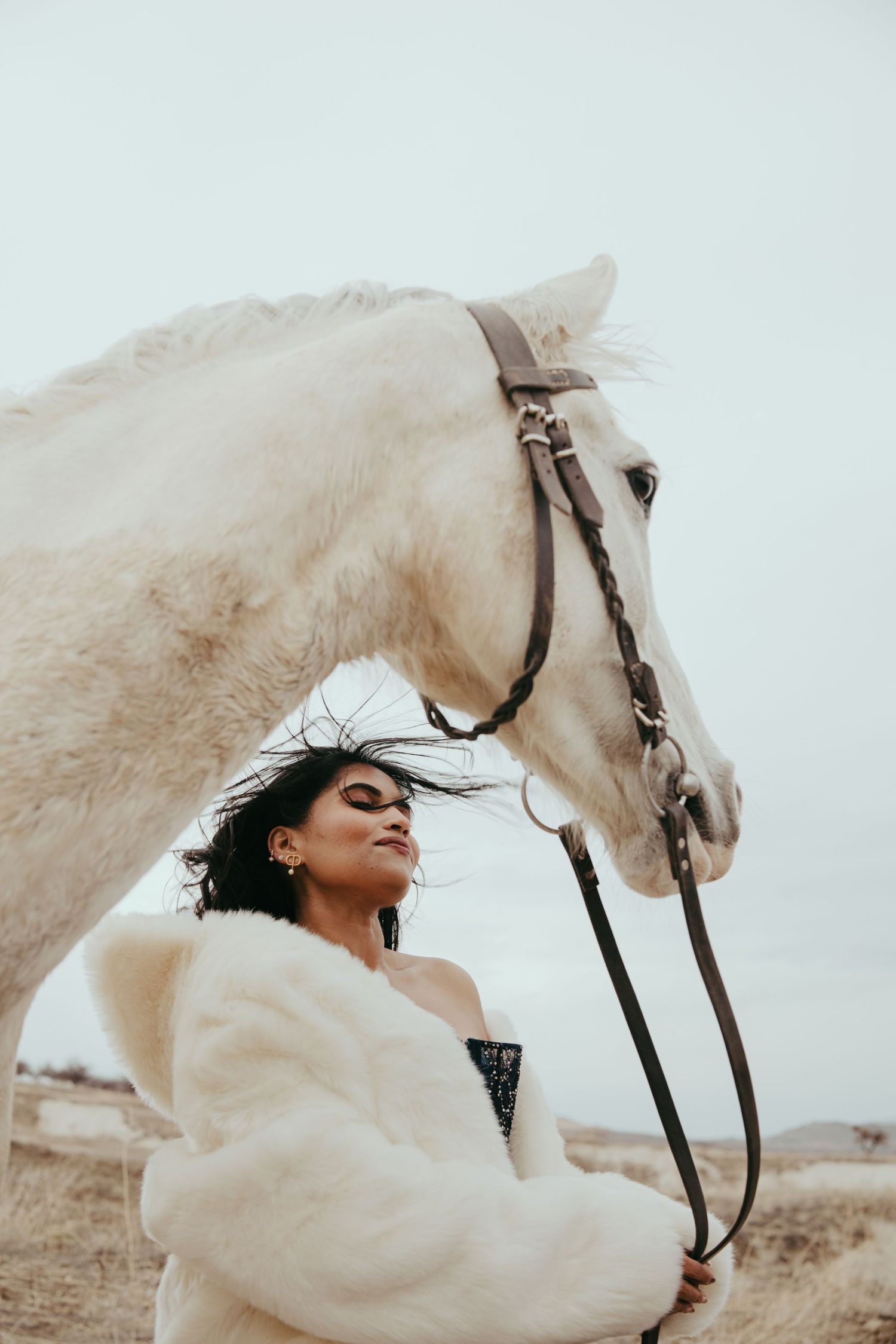 Woman in white fur coat stands beside a white horse with bridle in an open field.