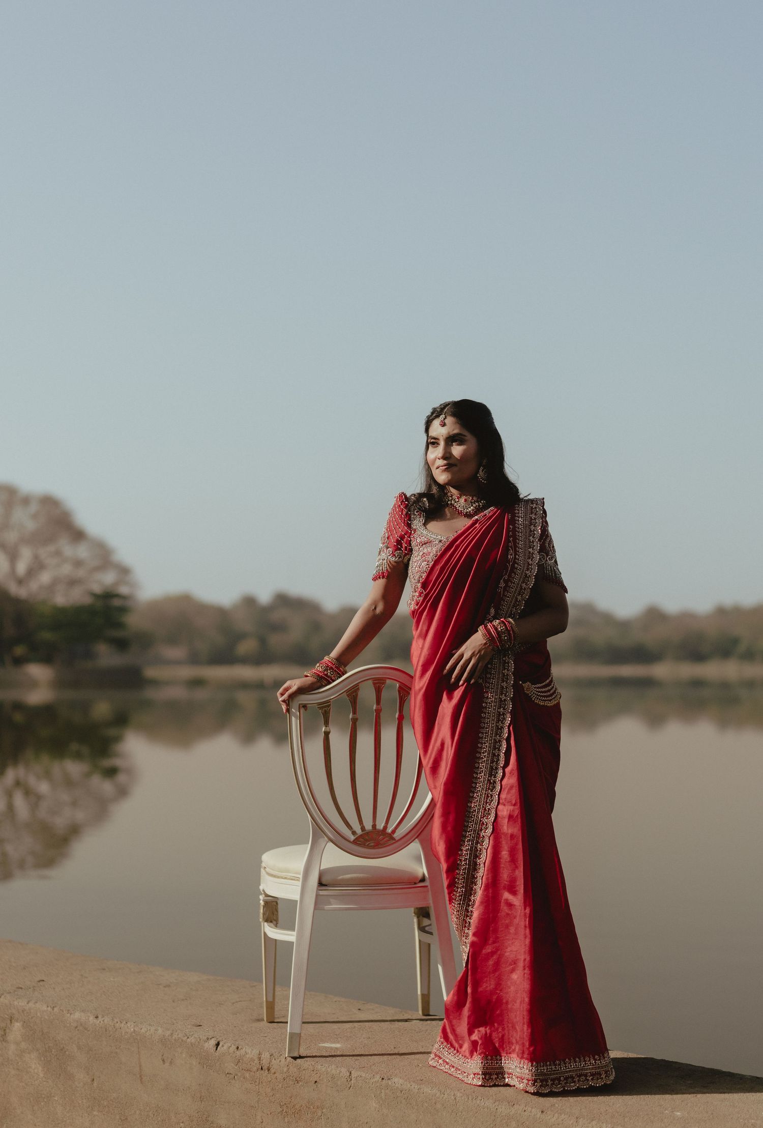 Bride in red saree standing by white chair near a serene lake.