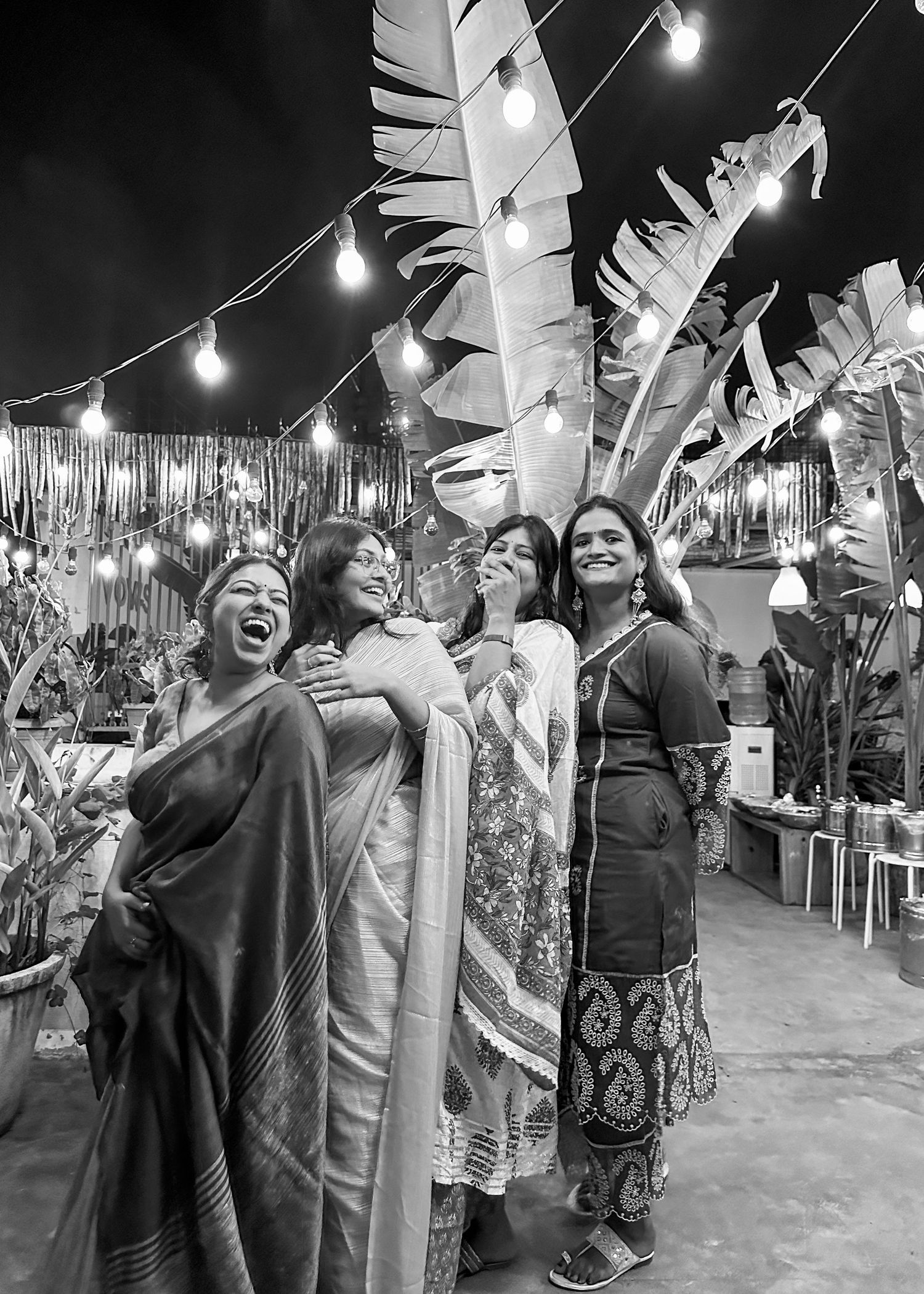 Four women in traditional South Asian attire laugh joyfully outdoors at a festive night event with string lights and tropical plants.