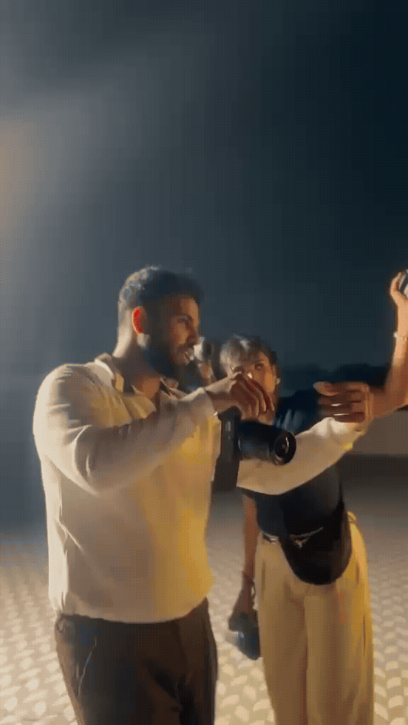 A bearded man in a cream shirt holds a camera while a person behind him gestures, lit by hazy backlight on a dark set.