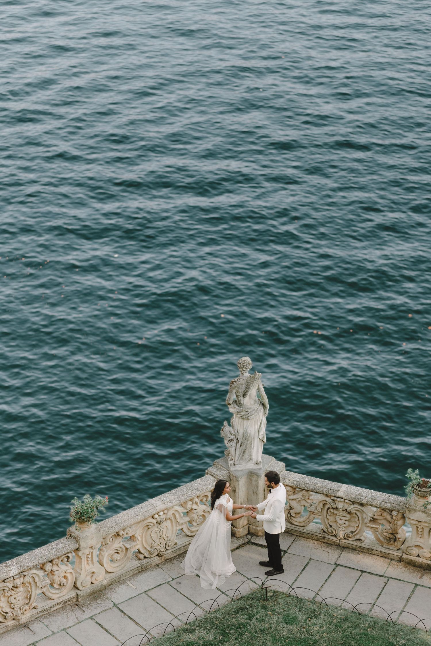 Couple holding hands on ornate terrace with classical statue overlooking turquoise lake waters.