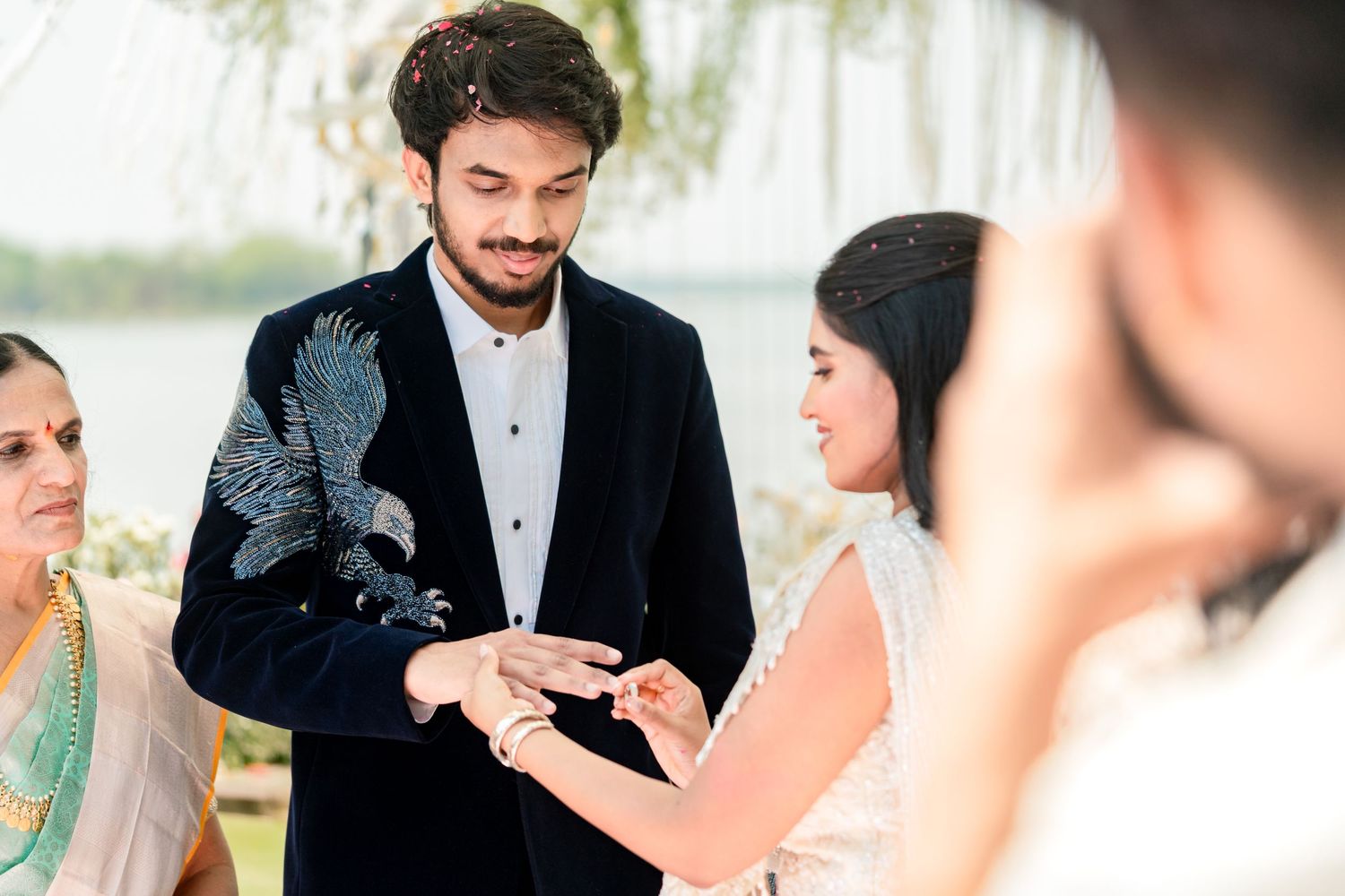 Groom in embroidered navy suit placing ring on brides finger during outdoor lakeside wedding ceremony.