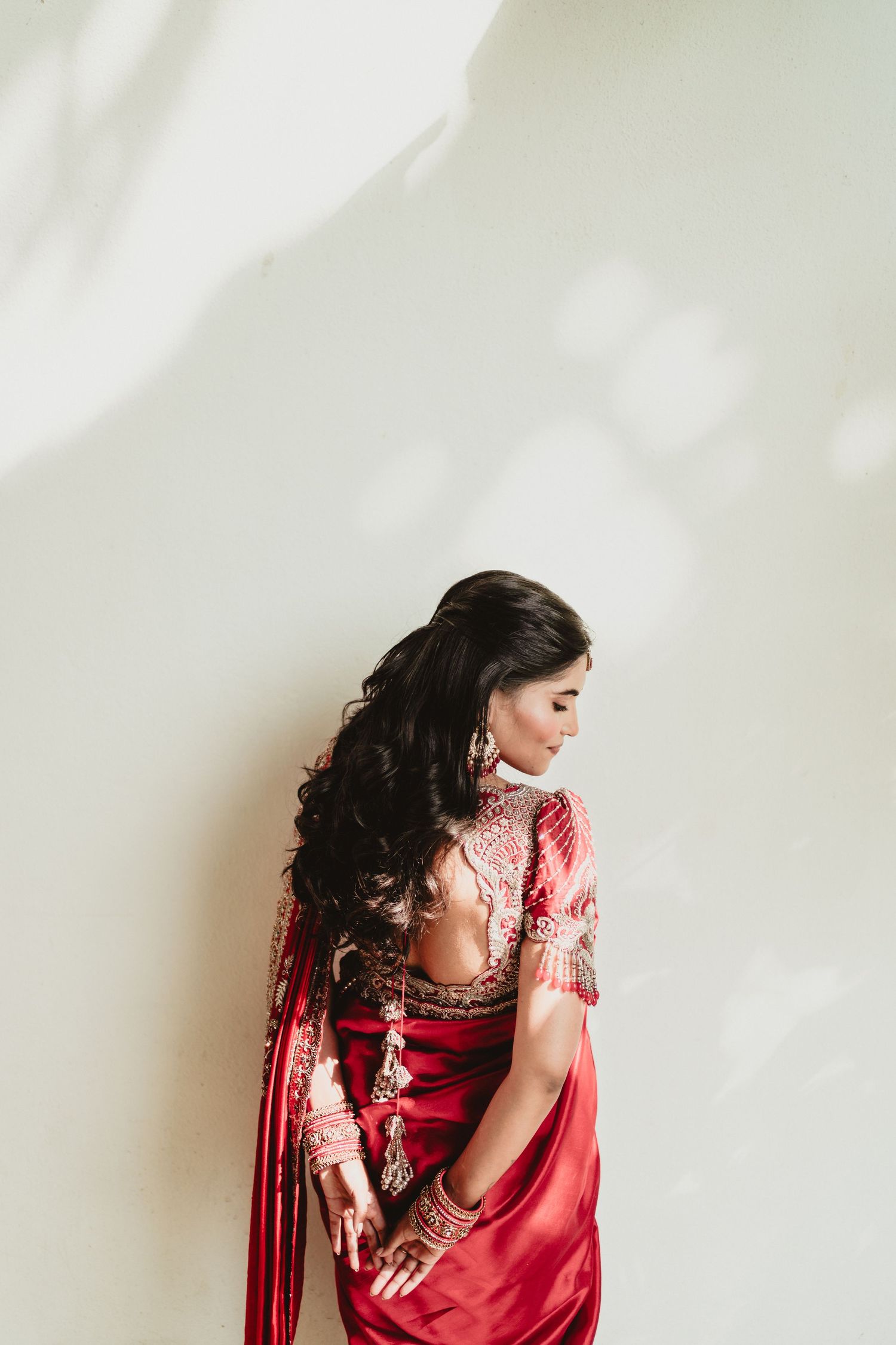 Indian bride in red embroidered saree with gold jewelry posing with back turned against white wall.