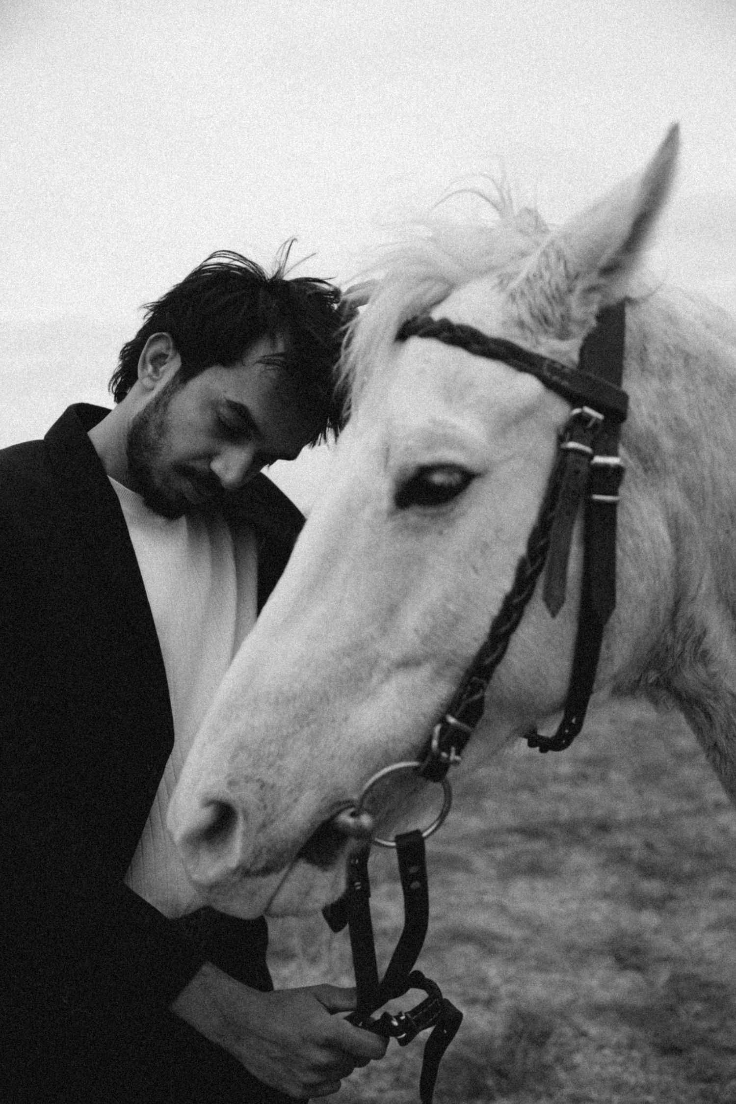A bearded man leans his head against a white horse wearing a bridle in a moody black and white portrait.