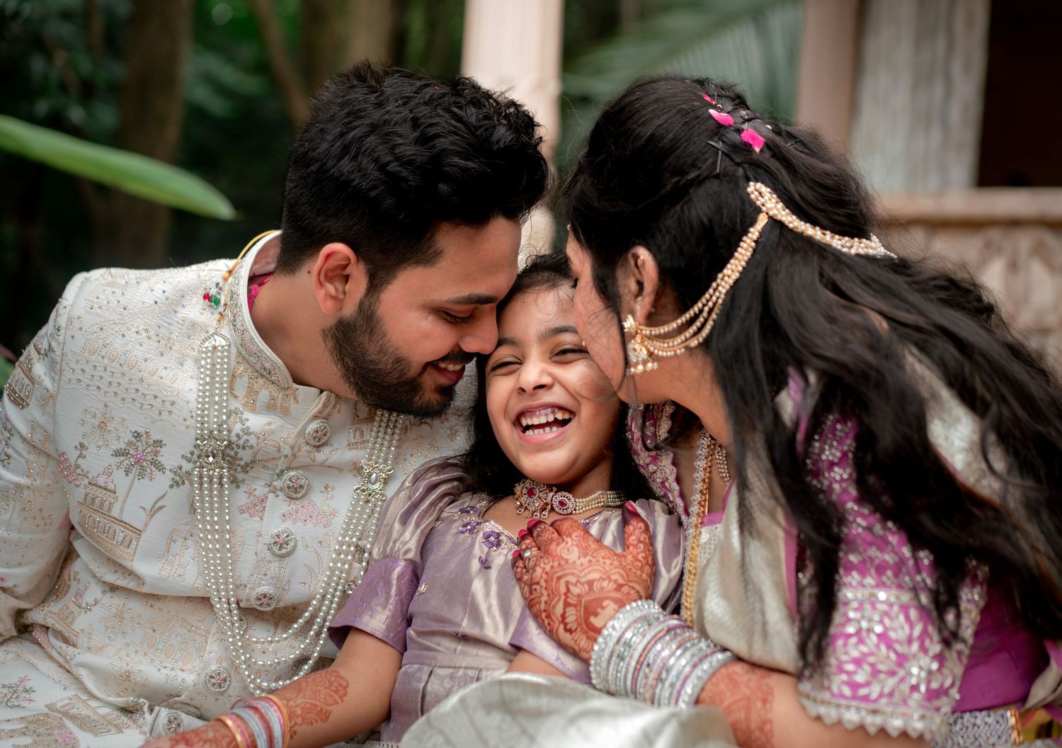 Indian couple in traditional wedding attire sharing a joyful moment with a young girl in a garden setting.
