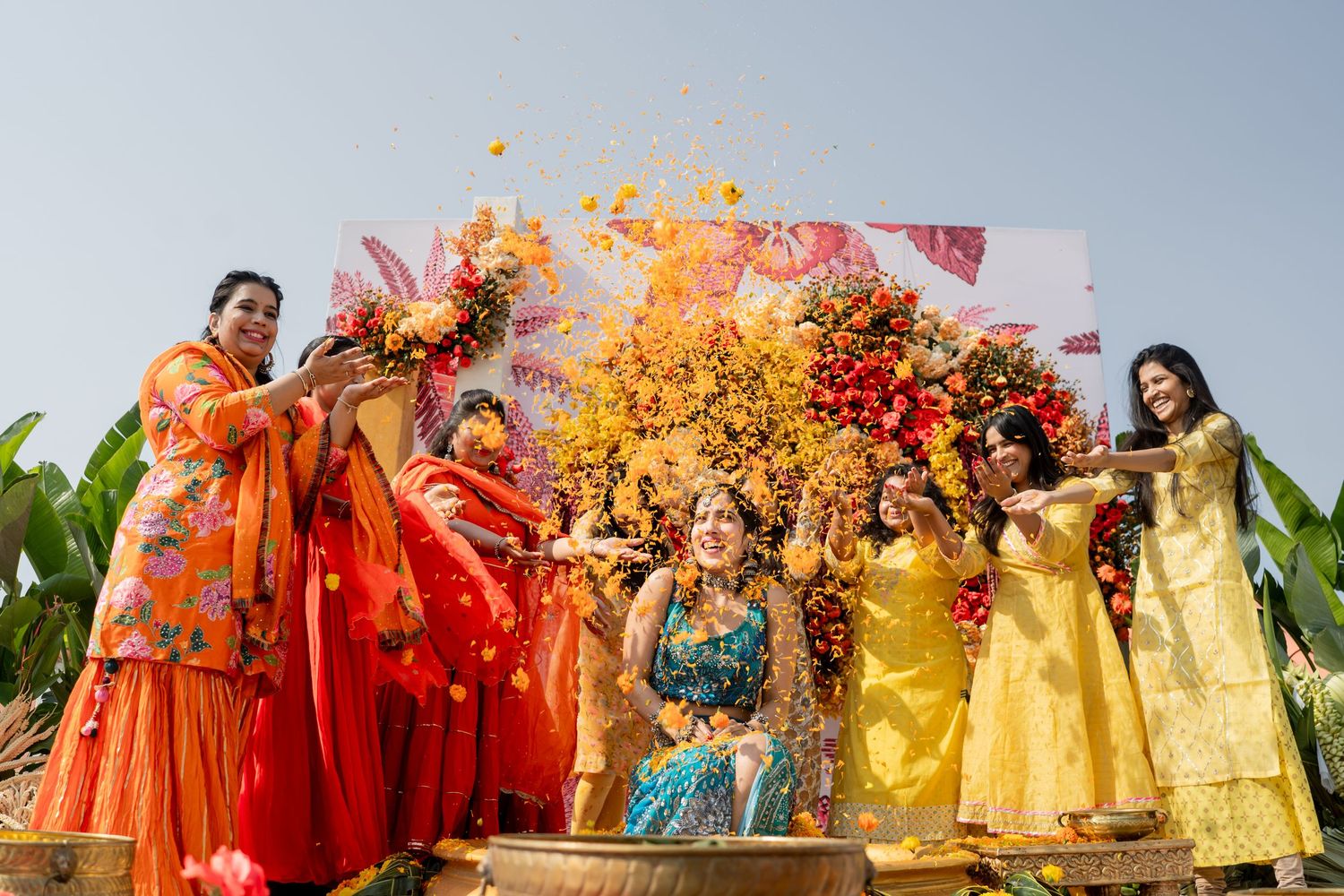 Women in colorful lehengas toss marigold flowers over a bride during a joyful Indian haldi ceremony.