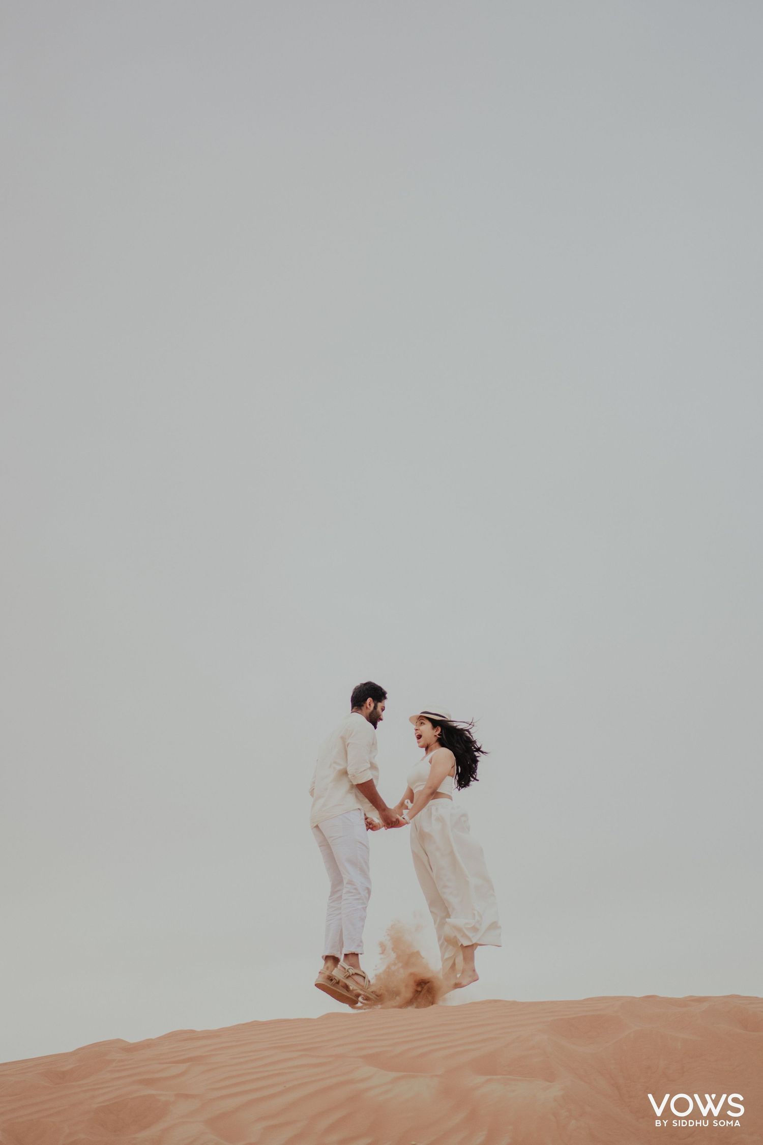 Couple in white outfits holding hands on a sandy desert dune, romantic pre-wedding shoot at golden hour.