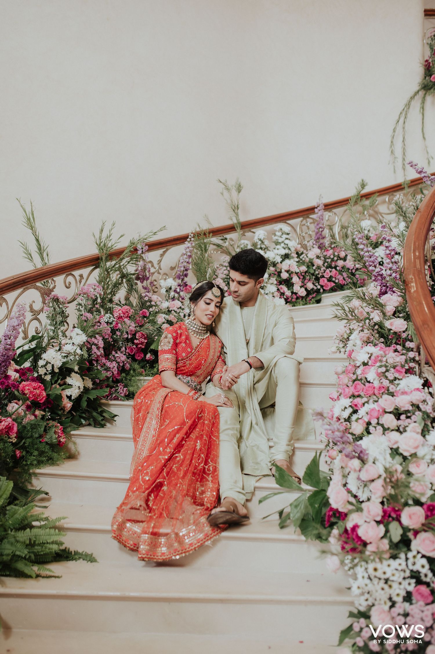 Indian bride in red lehenga and groom in ivory sherwani sit on flower-adorned stairs in a romantic wedding portrait.