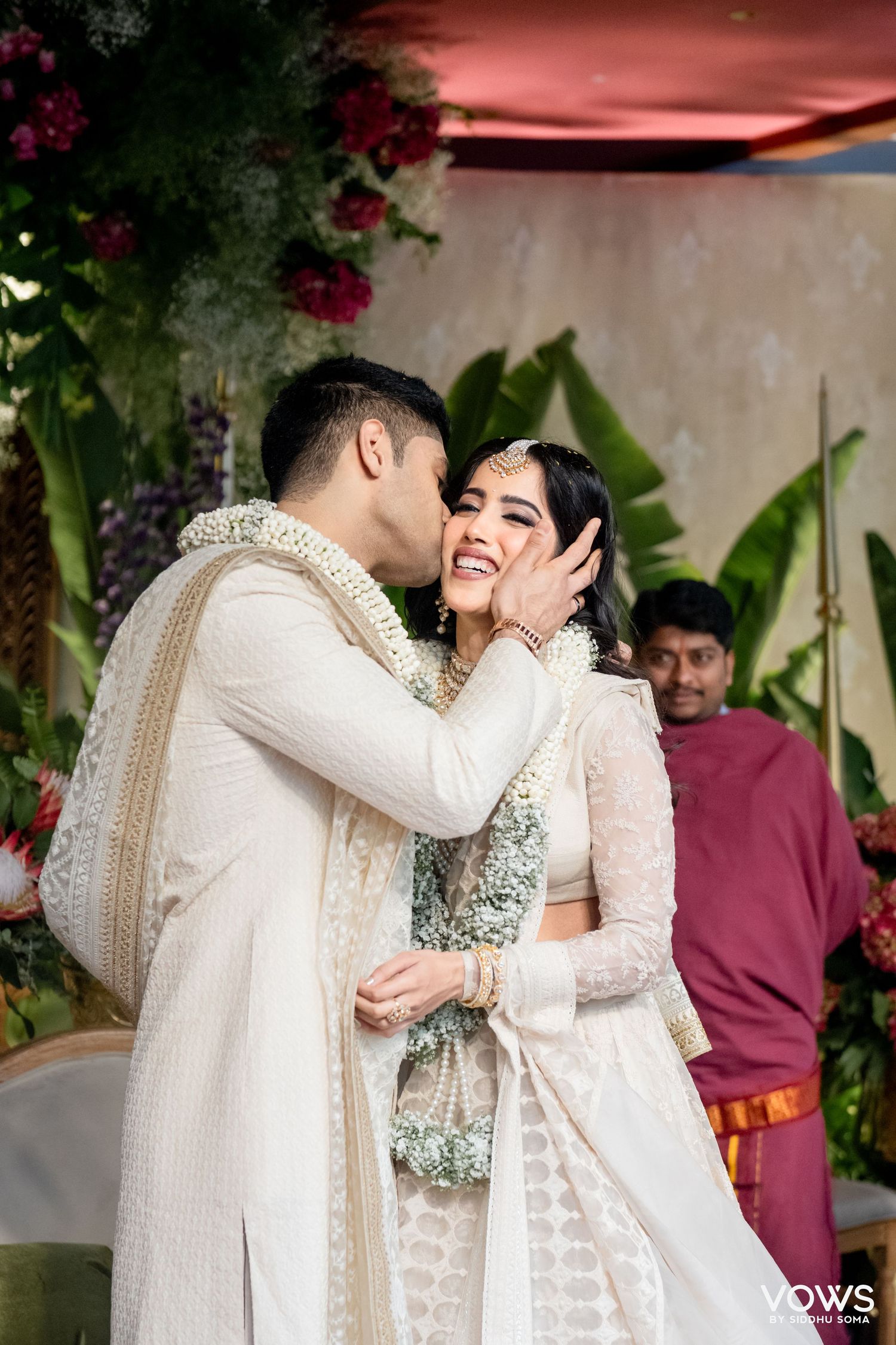 Groom in white sherwani kissing bride in white lehenga at Indian wedding ceremony with floral backdrop.