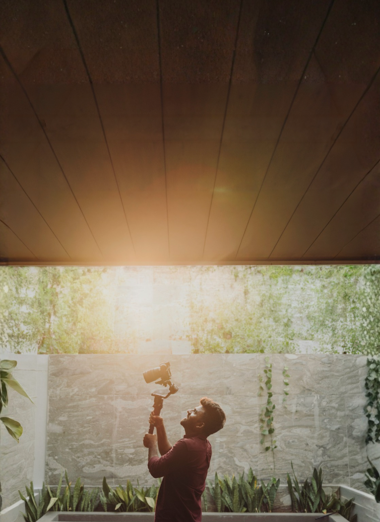 Man holding a camera on a gimbal outdoors, bathed in golden sunlight beneath a wooden ceiling.