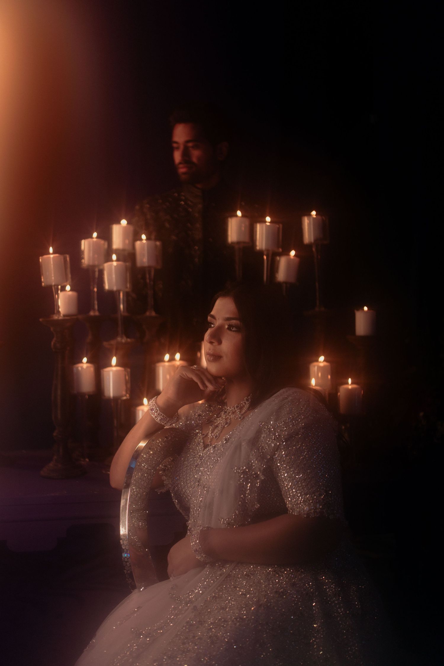 Bride in silver lehenga poses by candlelight with groom standing behind her in romantic dark setting.