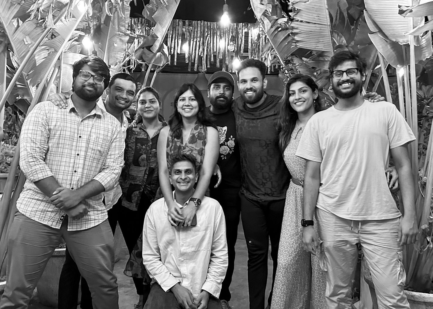 Black and white group photo of nine smiling friends posing together outdoors among tropical plants.