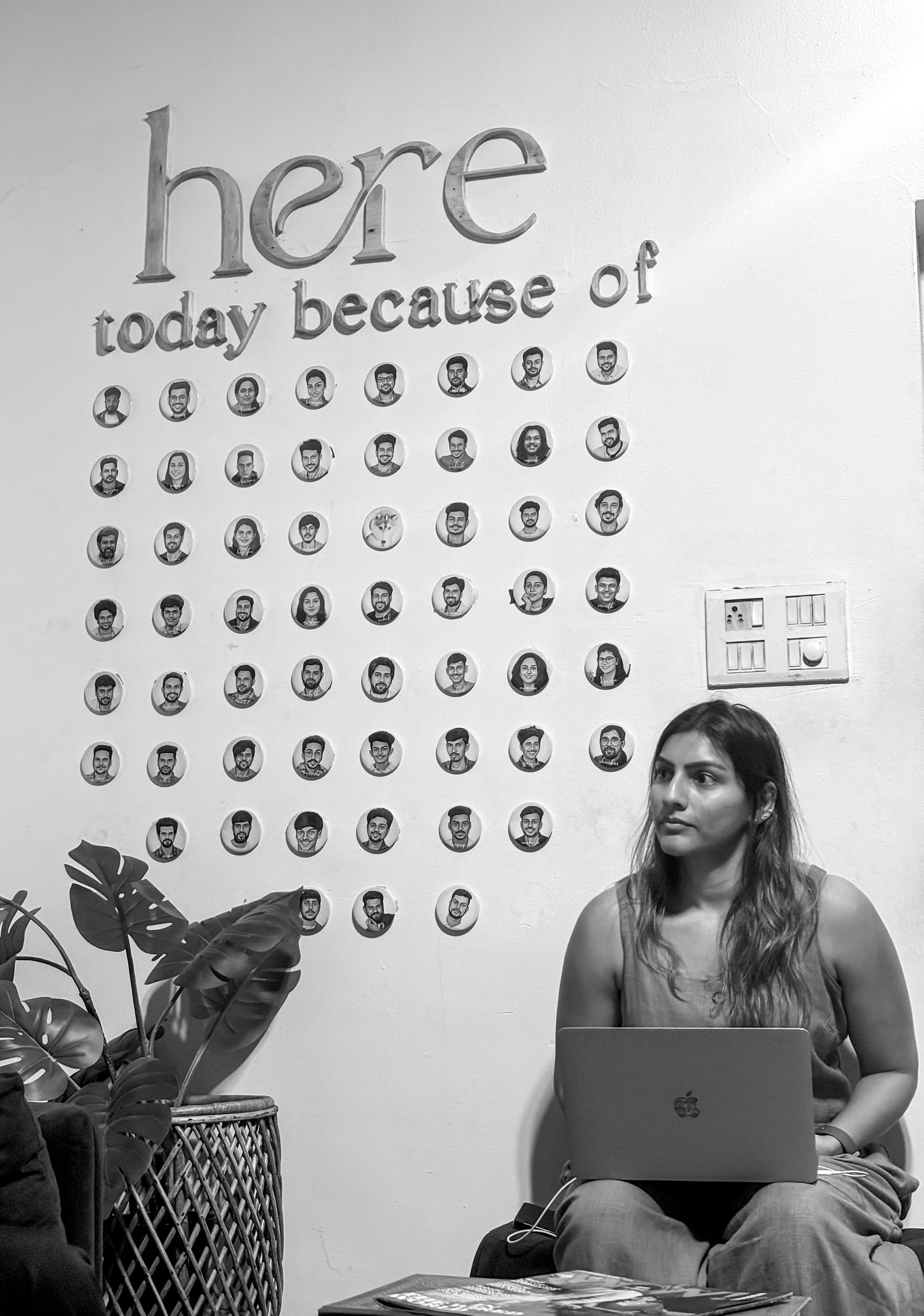 Woman sitting with laptop beneath a wall display reading 'here today because of' surrounded by circular portrait photos.
