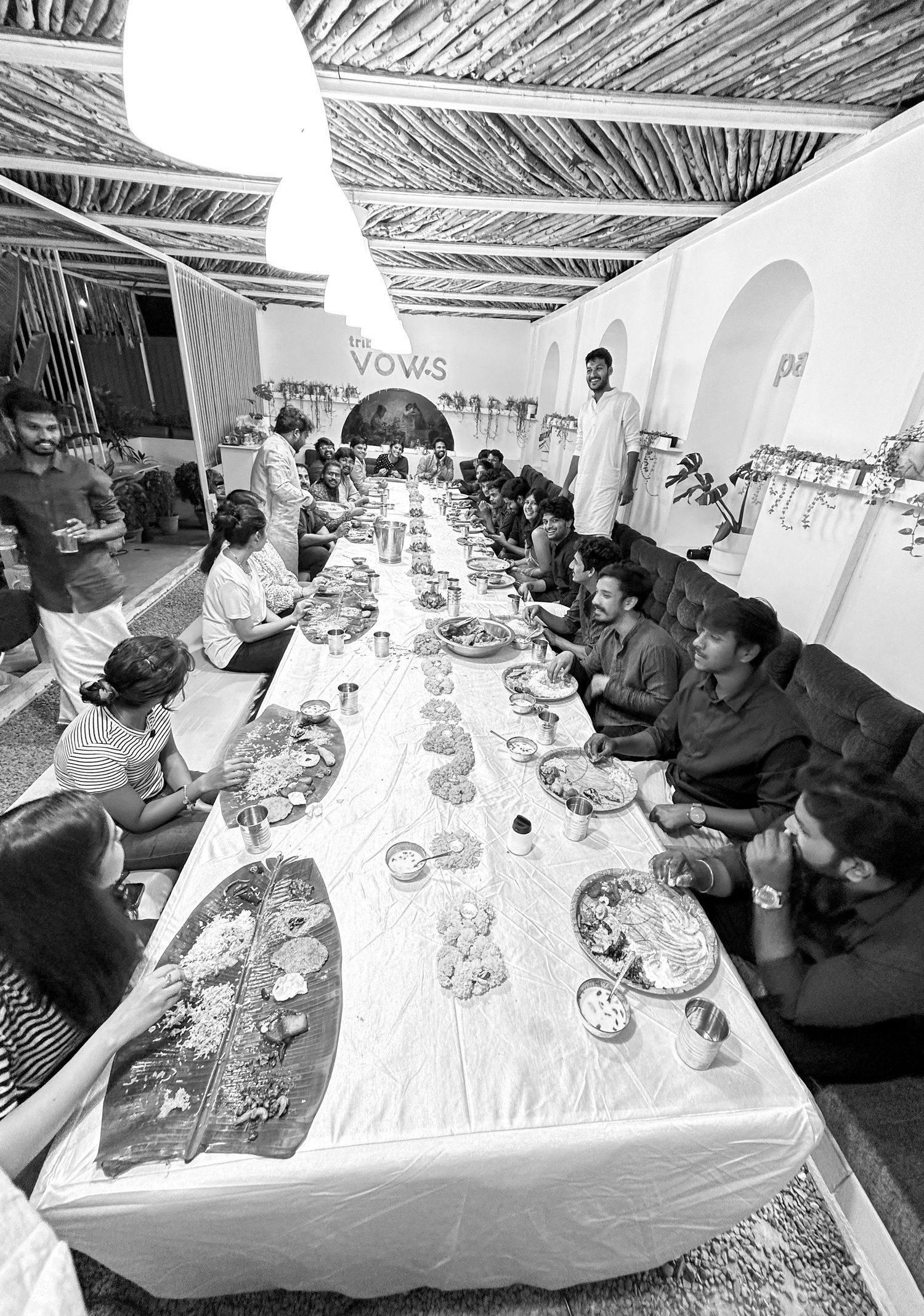 Black and white photo of guests dining at a long banquet table at Vows restaurant, featuring traditional Indian thali platters.