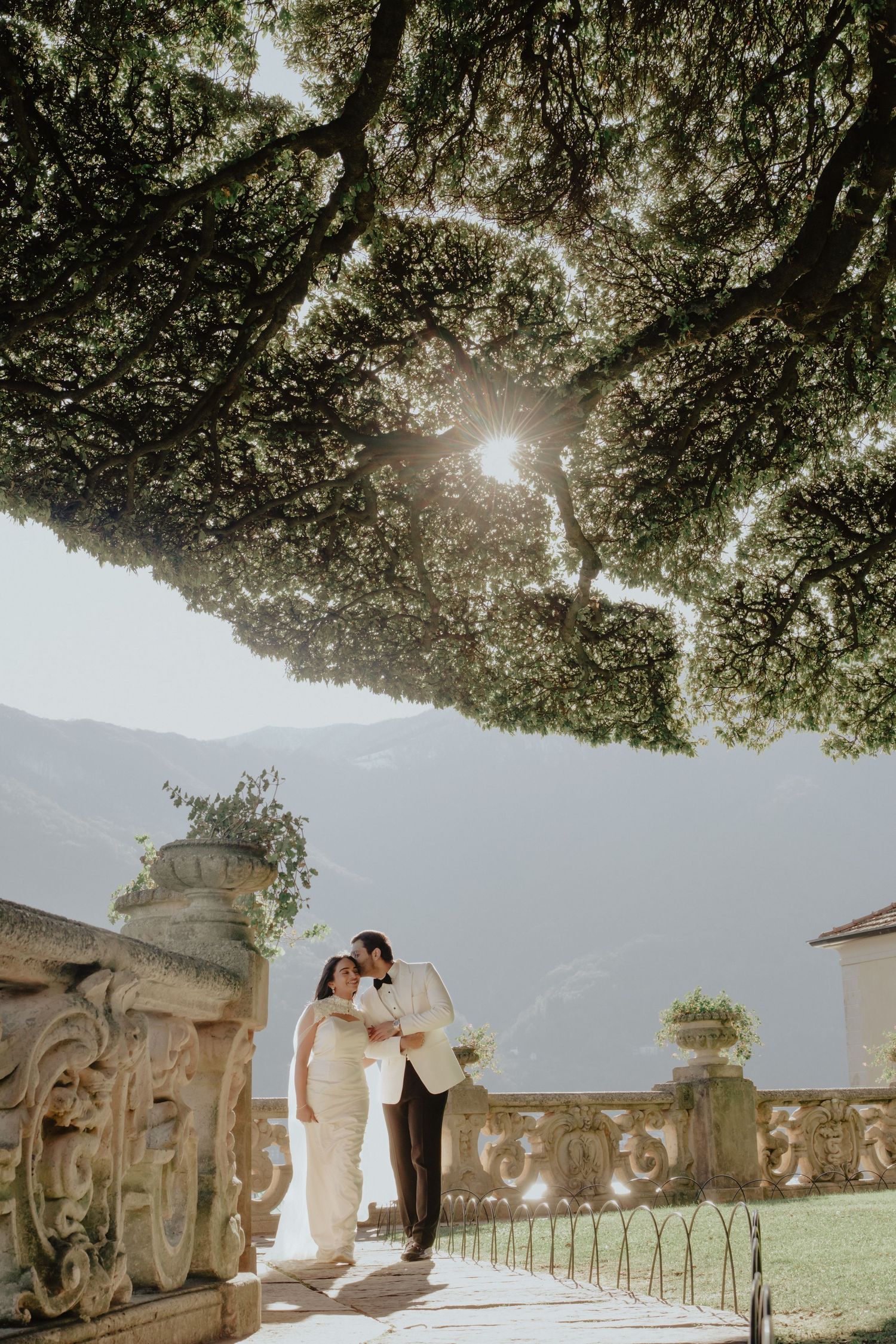 Bride and groom share a romantic moment under a sunlit tree at an elegant Italian villa wedding.