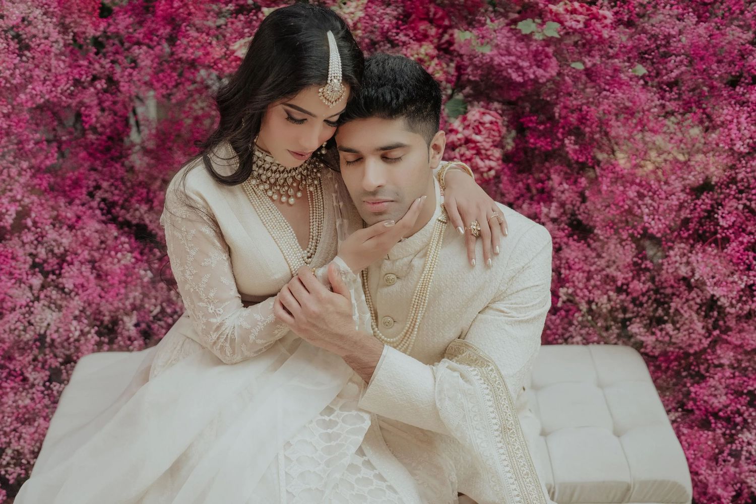 Couple in elegant white traditional attire posing romantically against vibrant pink bougainvillea floral backdrop.