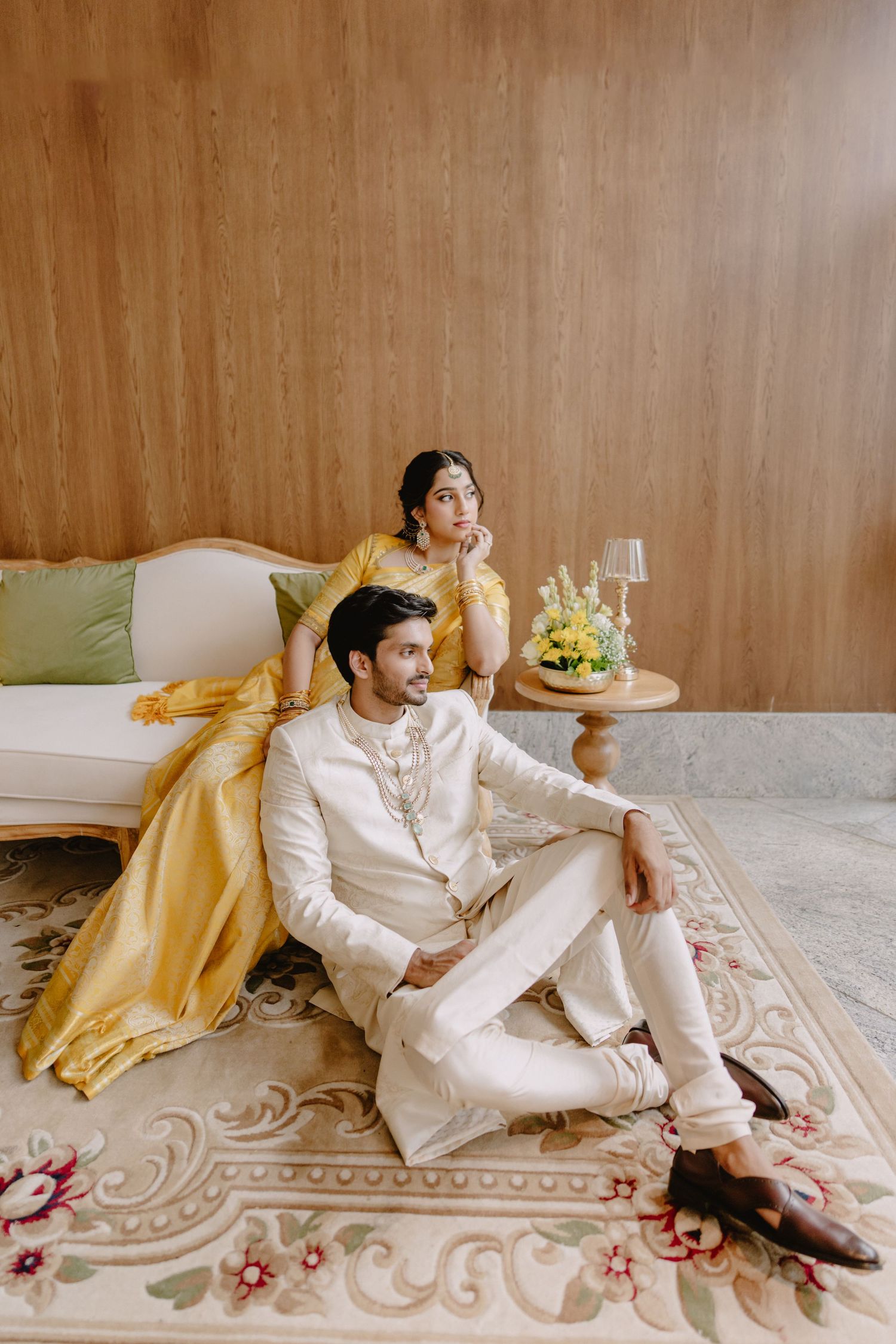 Indian couple in traditional attire, woman in yellow lehenga and man in white kurta, seated elegantly on a floral rug.