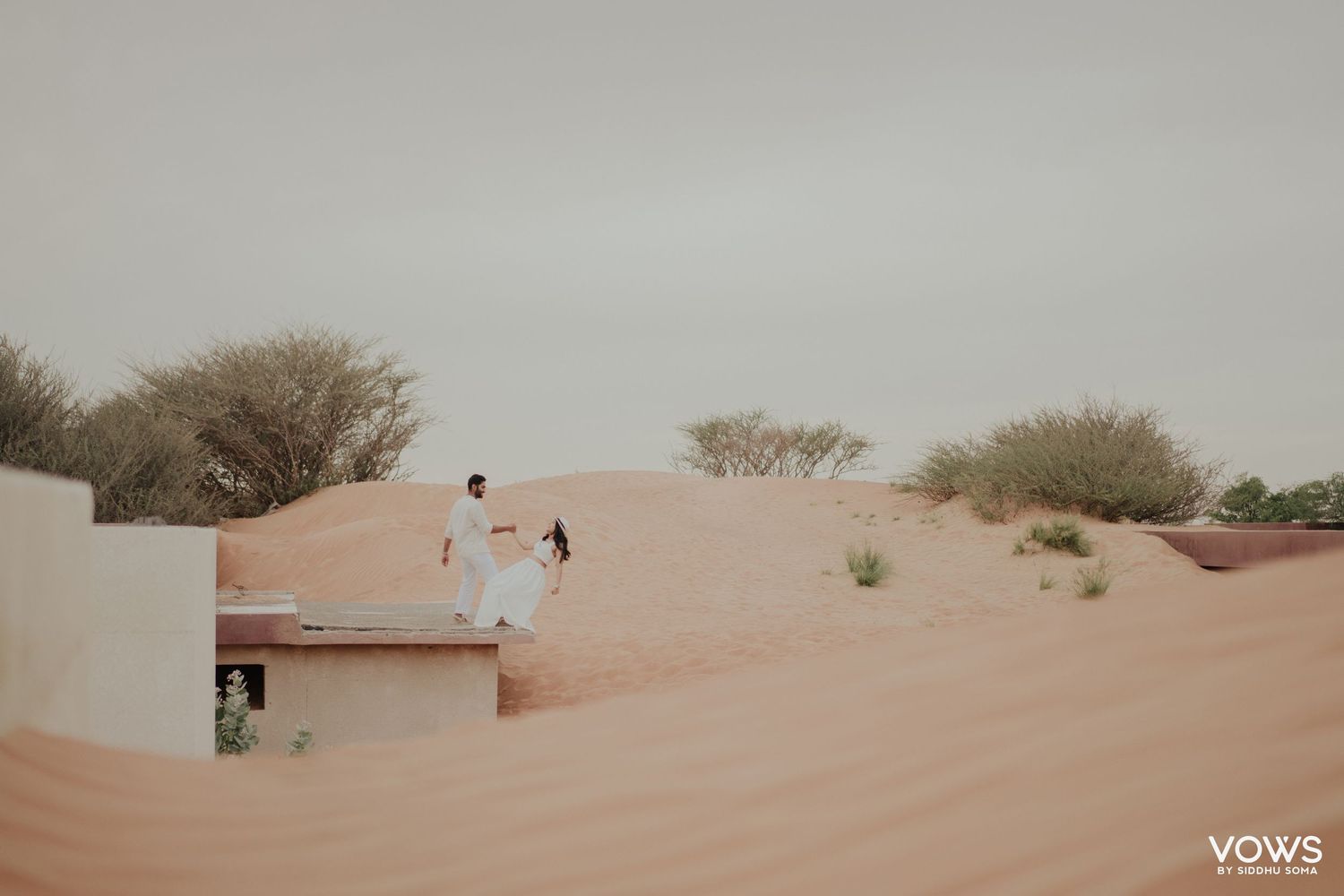 Couple in white attire walking on red sand dunes in a desert landscape with sparse green shrubs.