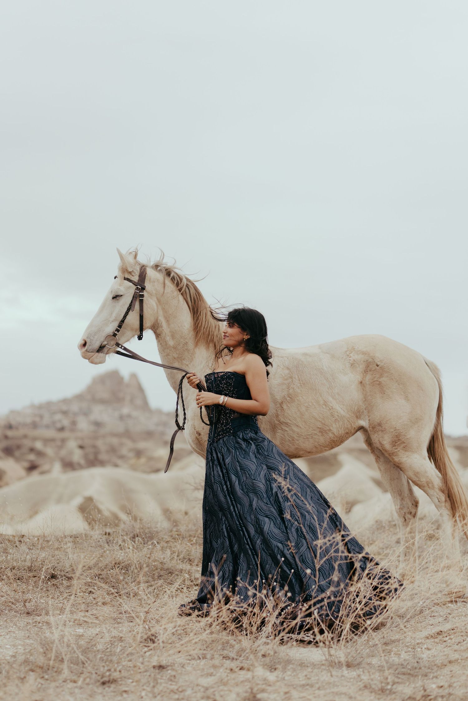Woman in black gown leading a white horse through arid landscape with rocky formations under overcast sky.