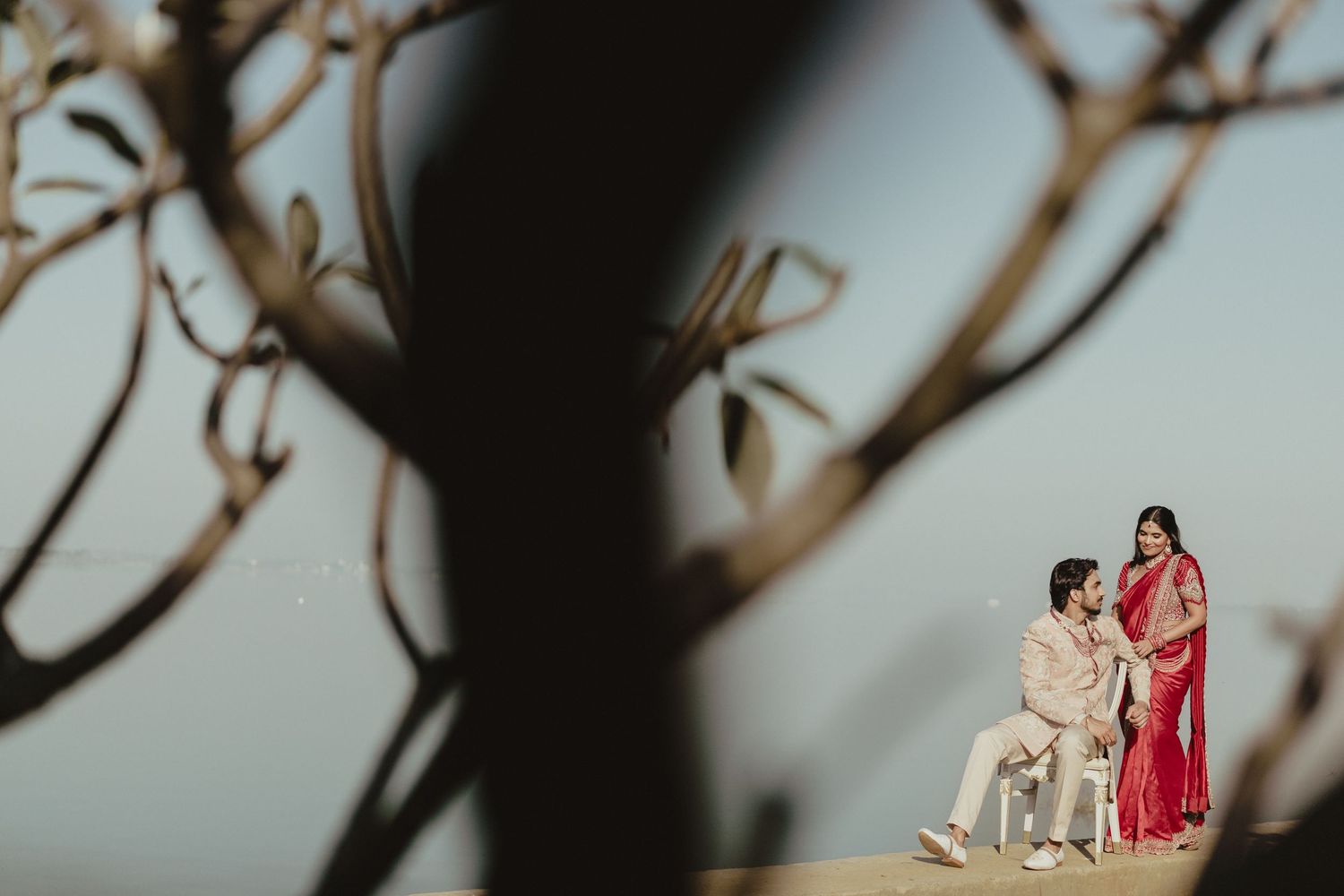 Couple in traditional Indian wedding attire standing on a hilltop framed by bare tree branches.