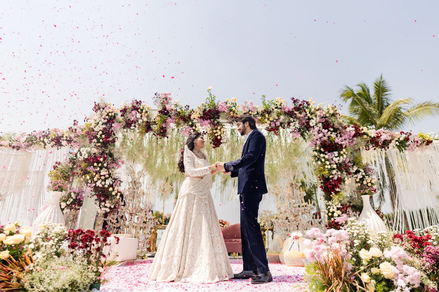 Couple exchanges vows under a stunning floral arch with pink blooms and rose petals at an outdoor Indian wedding ceremony.